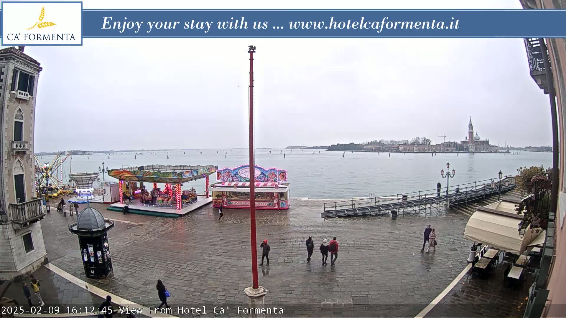 A wet, overcast day shows a plaza with a small carousel and several people walking near a body of water with a distant island.
