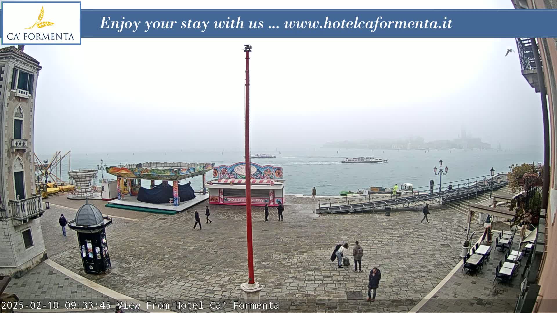 A foggy waterfront square with a small carnival, several people, and boats on the water.