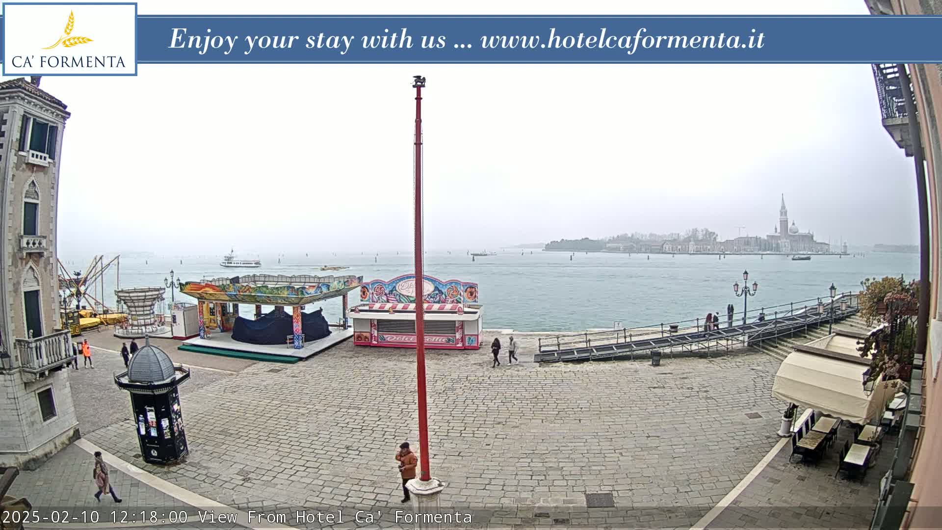 A waterfront plaza with carnival booths, a few people, and a distant view of a city across calm, gray water under an overcast sky.