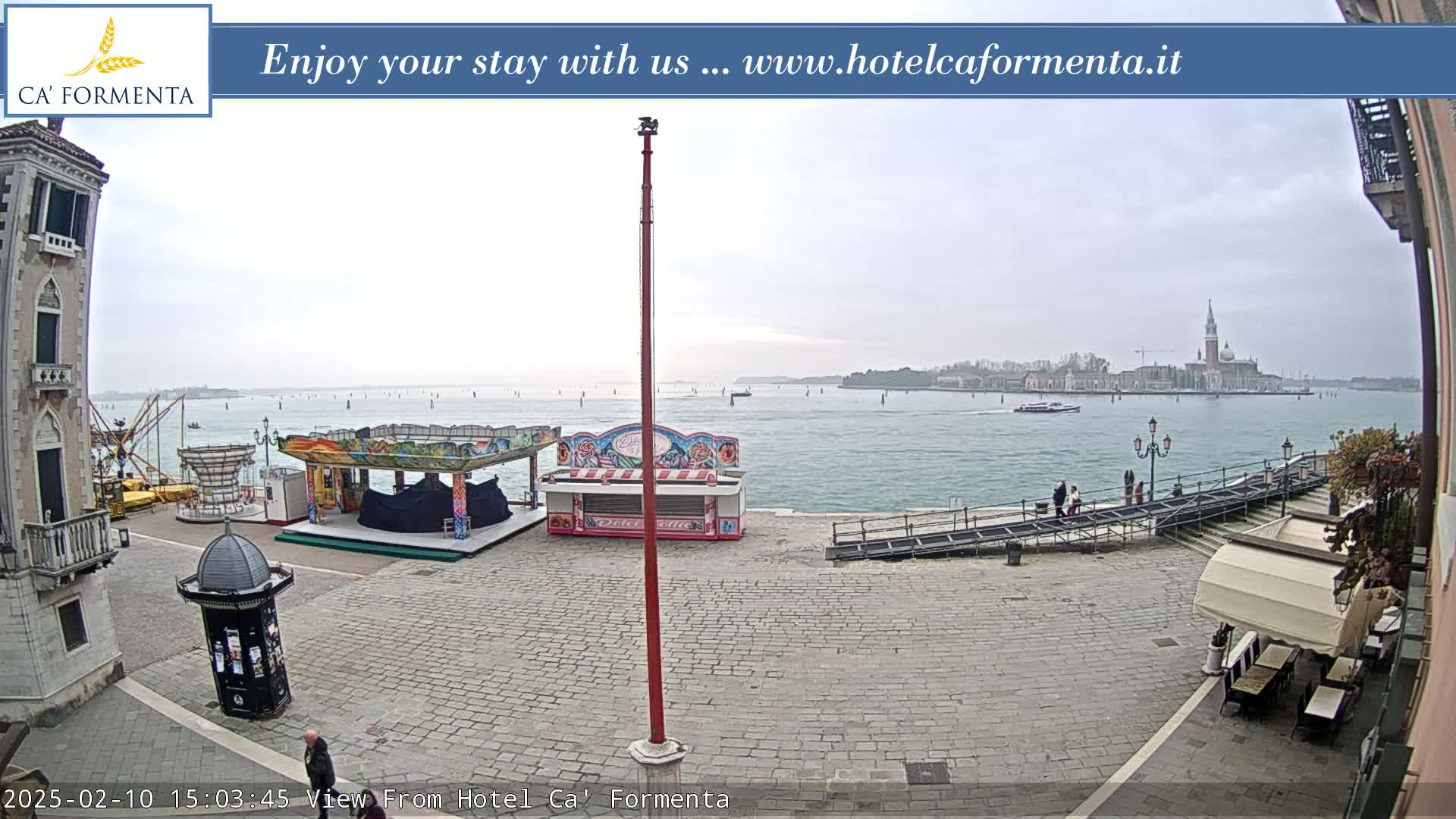 A paved plaza with carnival-style booths overlooks a calm body of water under an overcast sky, with a distant island visible in the background.