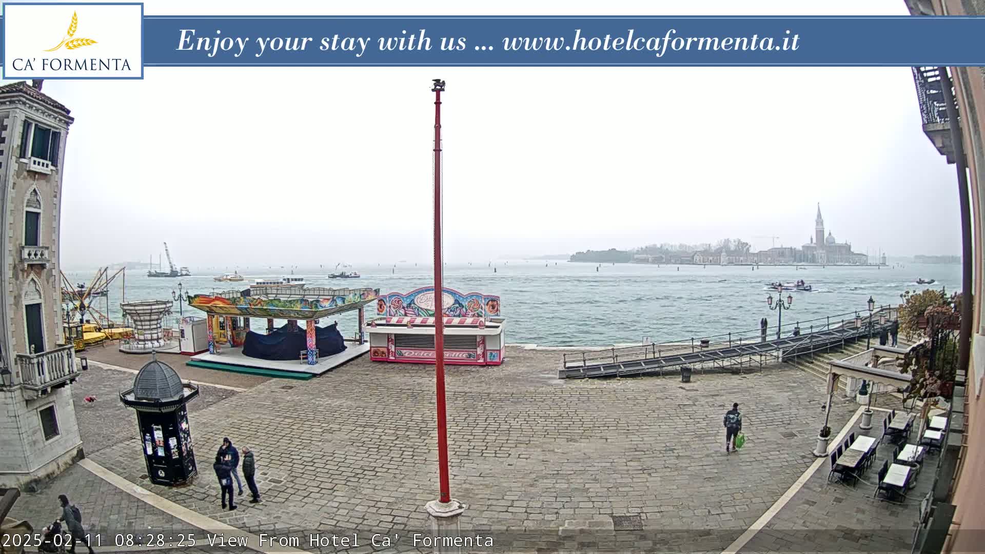 A stone plaza with a few people and carnival-style booths sits next to a calm body of water on an overcast day, with a distant city visible across the water.