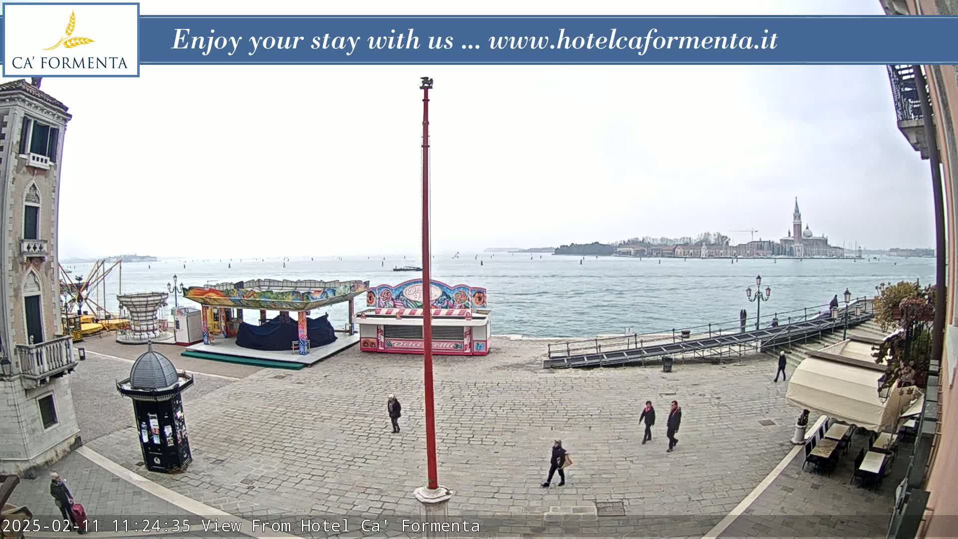 A paved square overlooks a calm body of water with distant buildings and a few people walking, under an overcast sky.