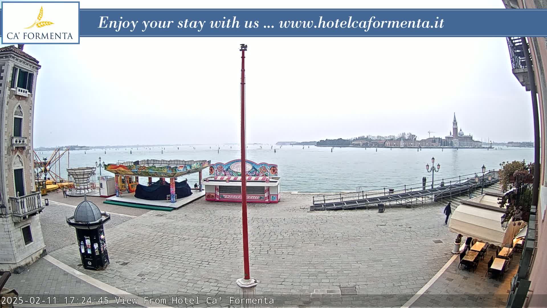 A waterfront plaza with carnival rides and an outdoor cafe overlooks a calm body of water on an overcast day, with a distant cityscape visible across the water.