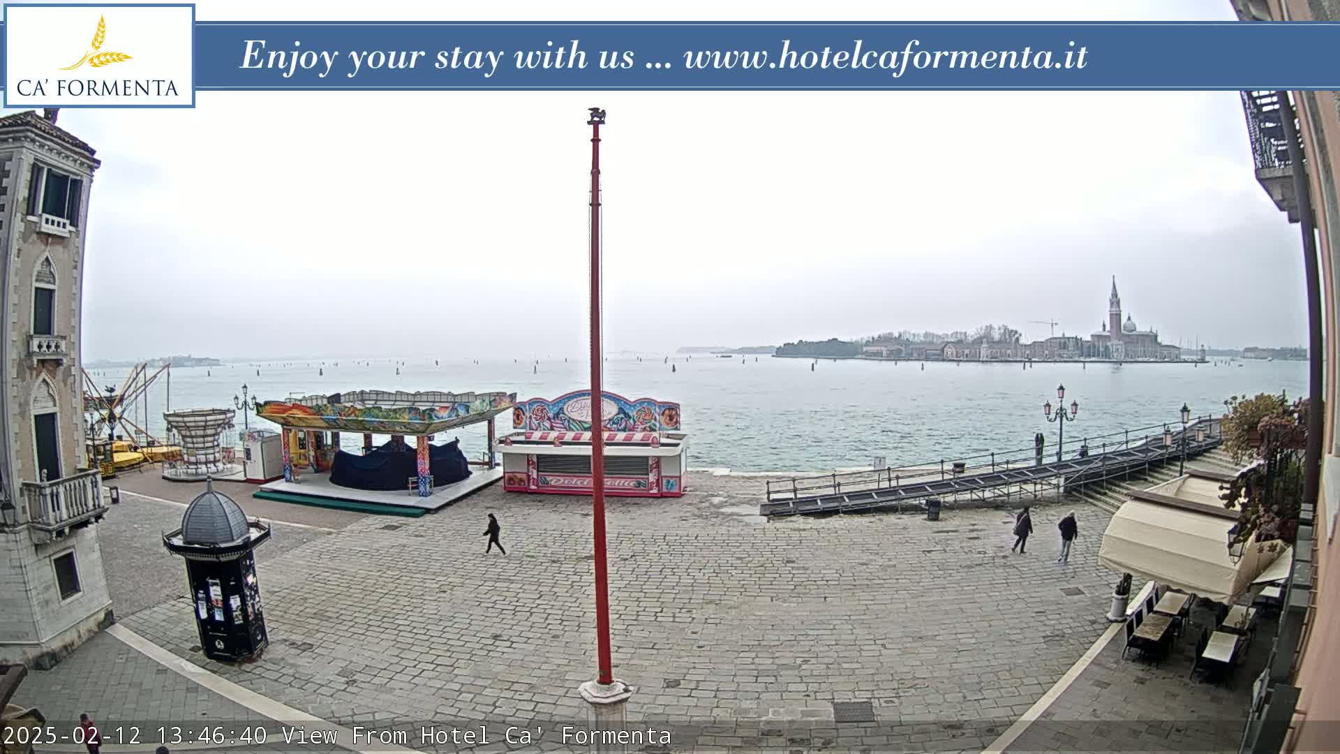 A paved square near a body of water with a carnival-like setup, featuring several small kiosks and rides under an overcast sky, overlooks a distant island with buildings.