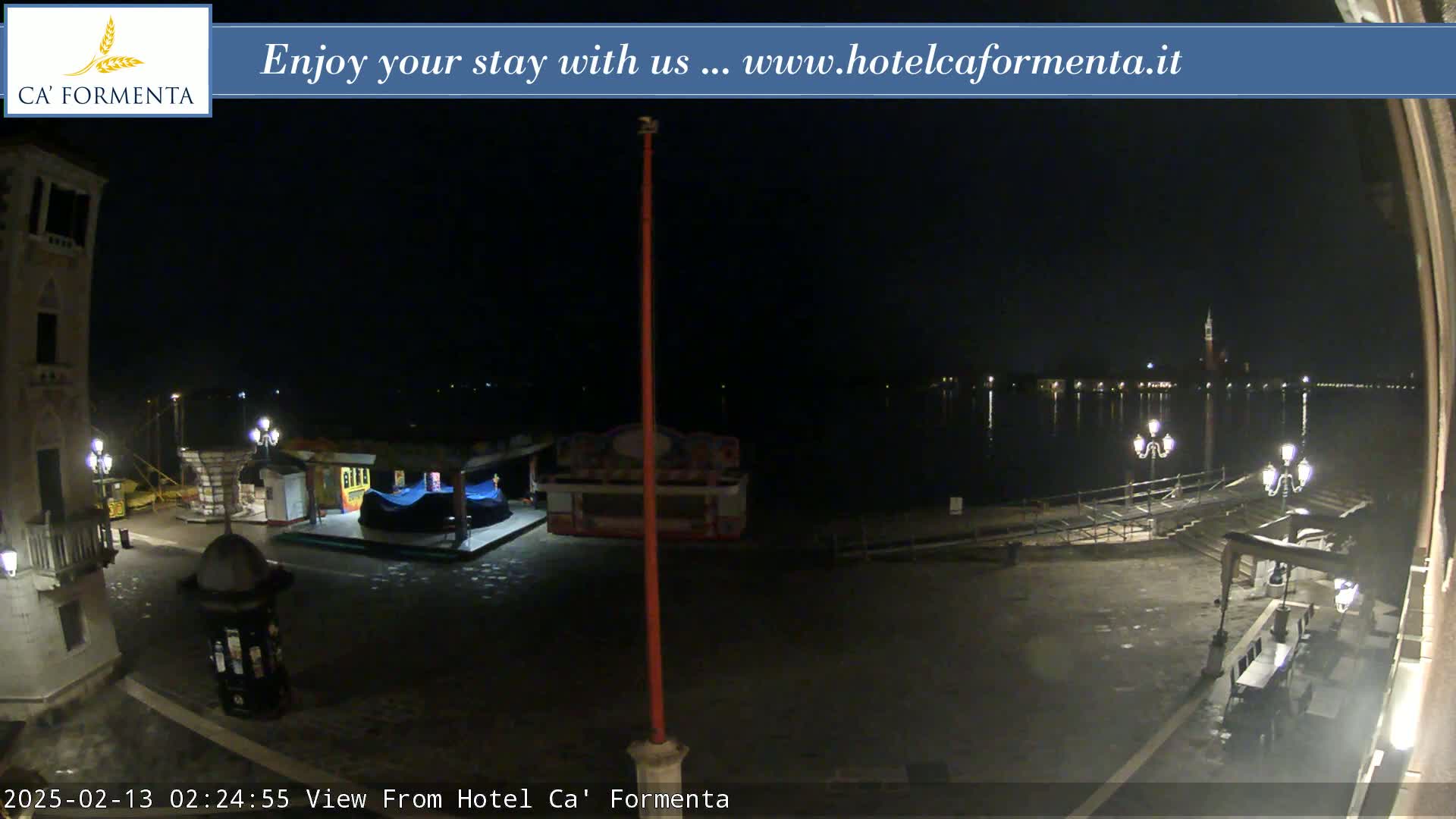 A nighttime view of a waterfront plaza with a calm body of water, some small structures, and streetlights.
