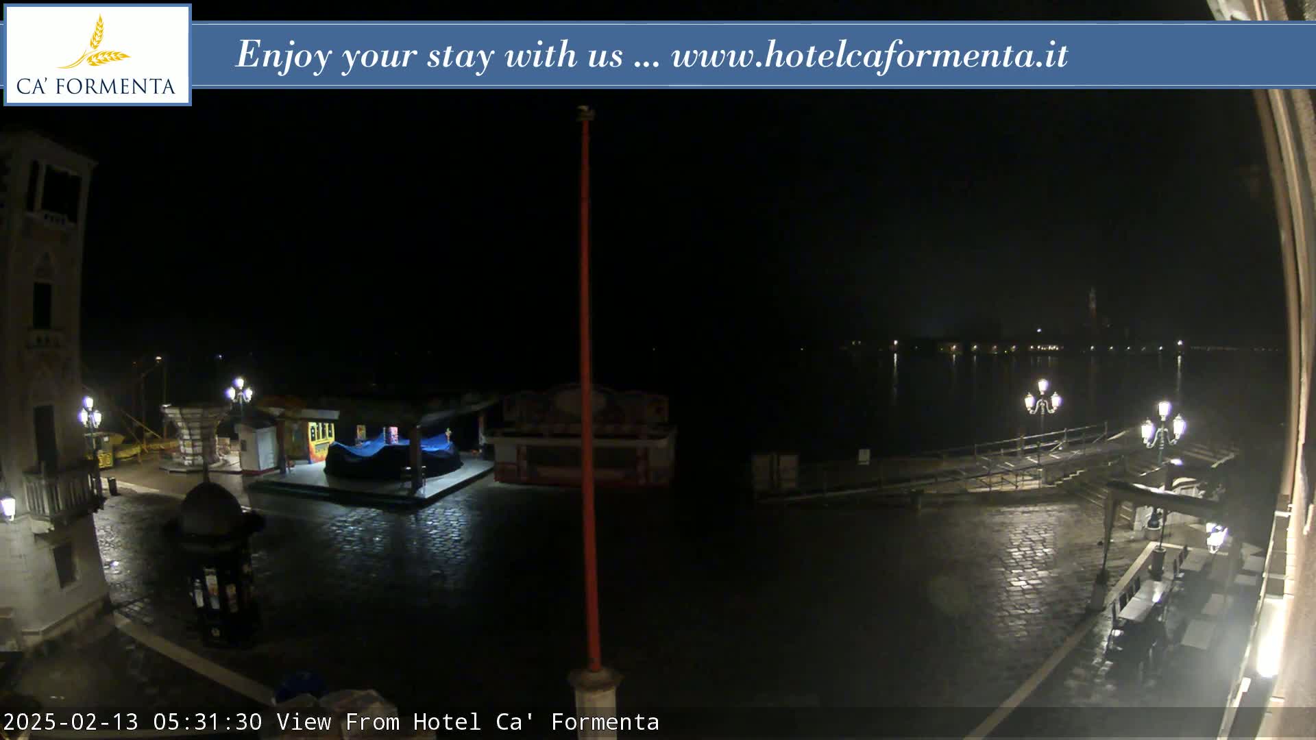 A nighttime view of a waterfront plaza shows a dark body of water, wet cobblestone streets, and a few illuminated structures.