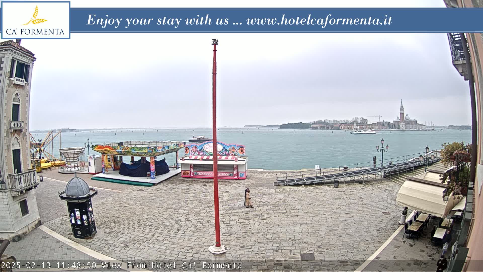 A waterfront plaza with carnival-style booths and a distant view of a city across a calm, overcast body of water.