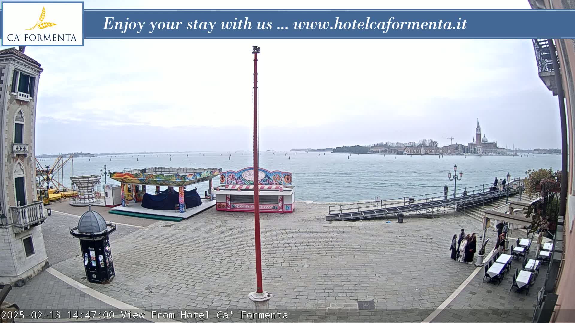 A paved waterfront square with carnival rides, a calm body of water, and a distant island on an overcast day.