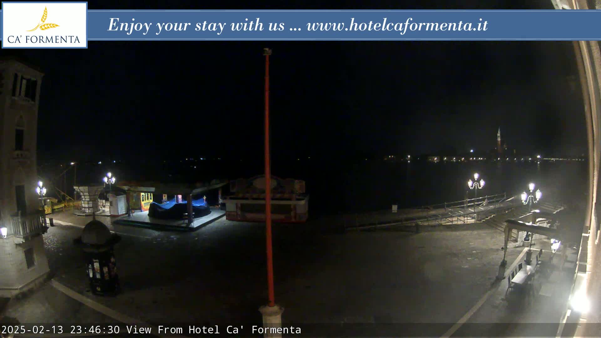 A nighttime view of a waterfront plaza with a few small kiosks and a calm body of water showing distant lights across it.