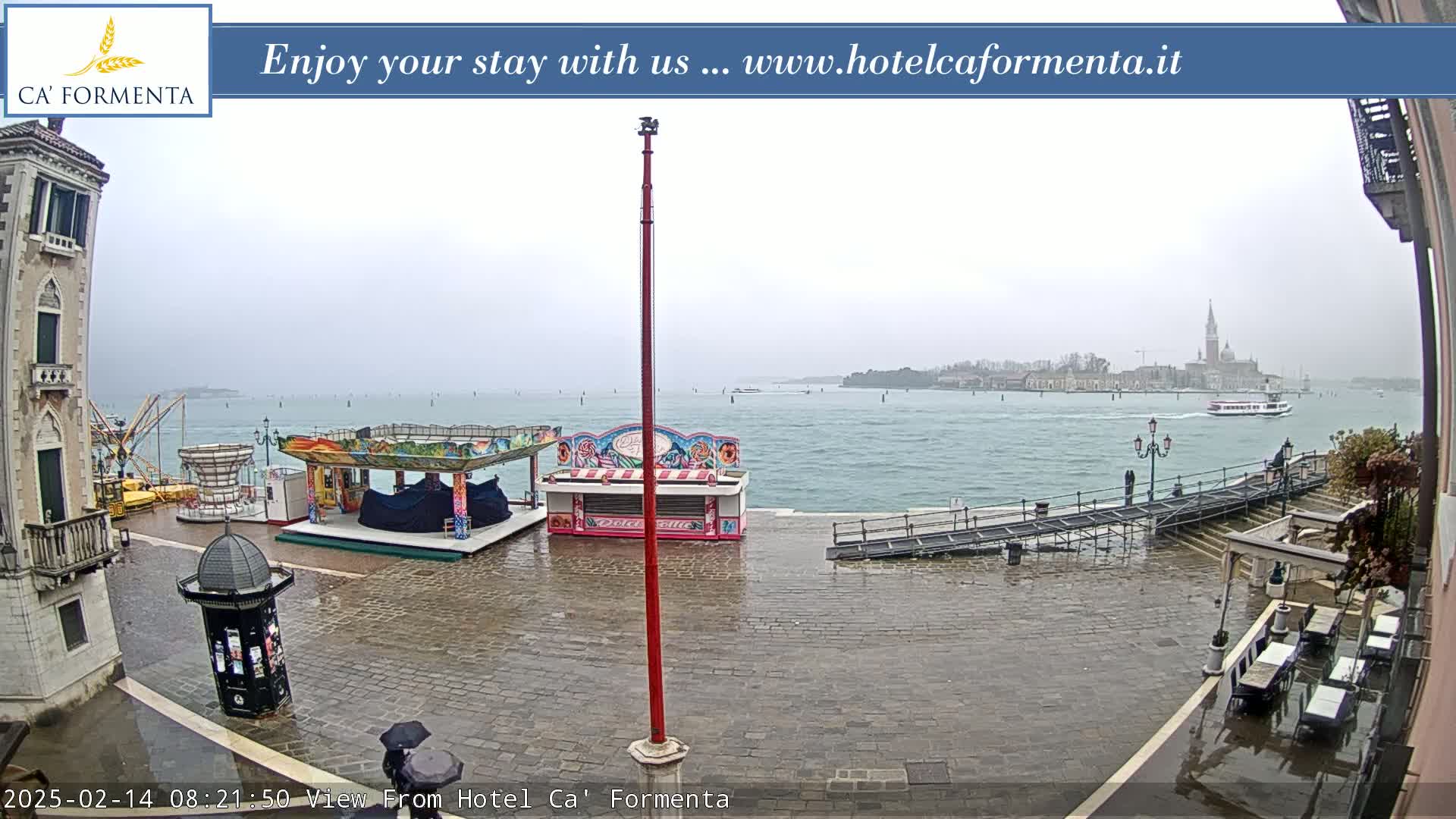 A rainy waterfront view shows a cobblestone plaza with carnival-style food stands and a distant view of a city across the water.