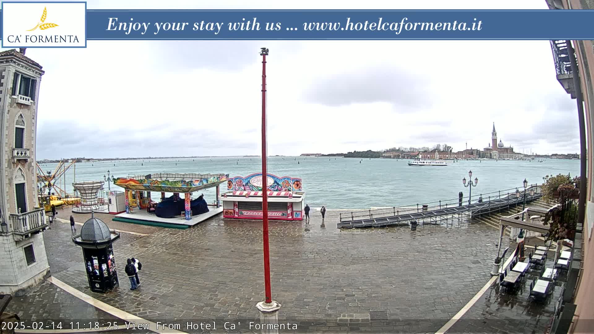 A wet cobblestone plaza with carnival rides, a body of water, and a distant cityscape under an overcast sky.