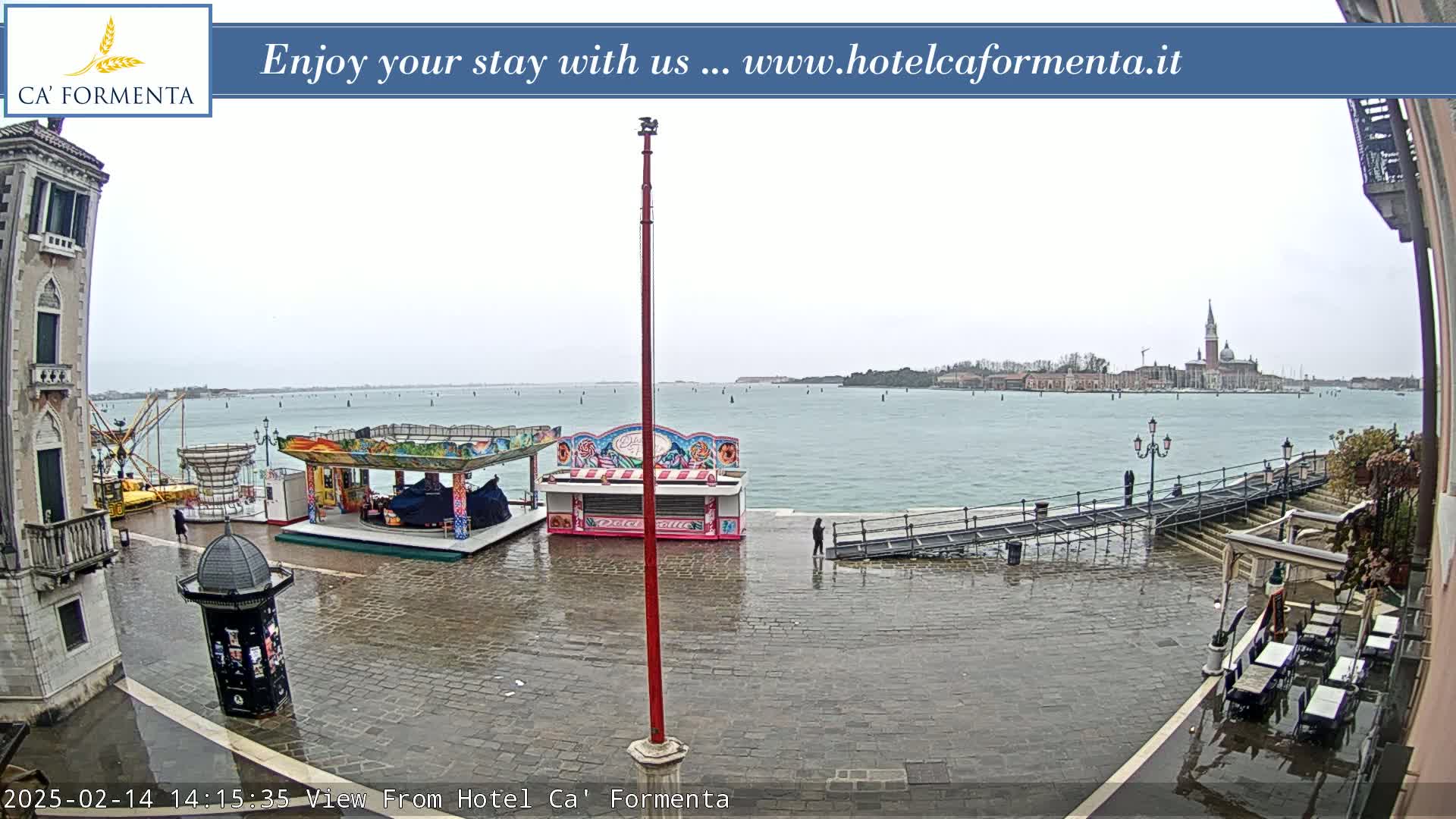 A wet, overcast day in Venice shows a waterfront plaza with a small amusement park, a distant view of the city across the water, and outdoor seating at a nearby restaurant.