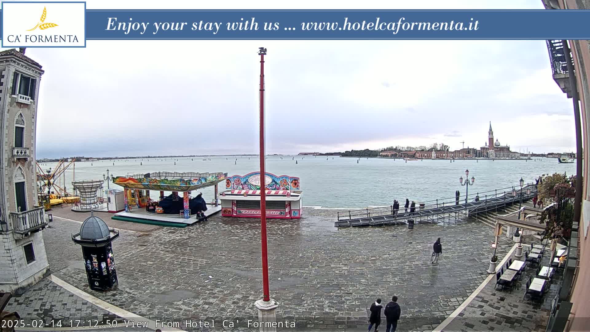 A mostly-empty cobblestone plaza with carnival rides and outdoor seating overlooks a calm body of water under an overcast sky, with a distant cityscape visible across the water.