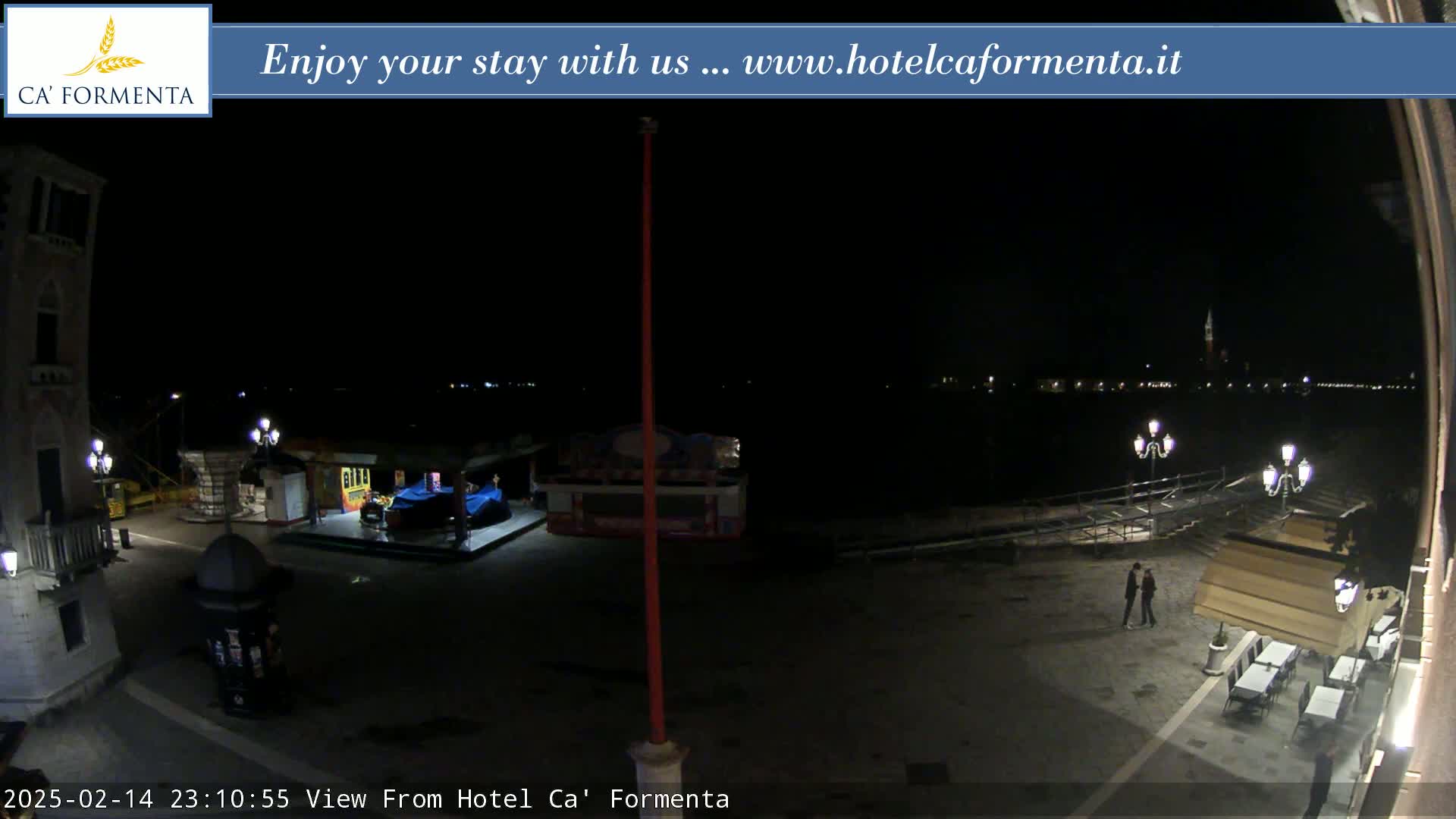 A nighttime view of a waterfront plaza shows a few people near outdoor seating, a small carnival ride, and a dark body of water with distant lights.