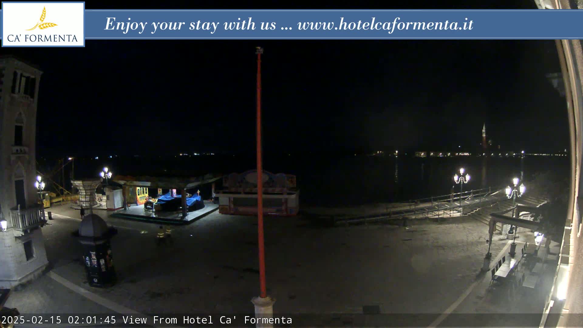 A nighttime view of a waterfront plaza with a few lights, a small structure, and calm water in the background.