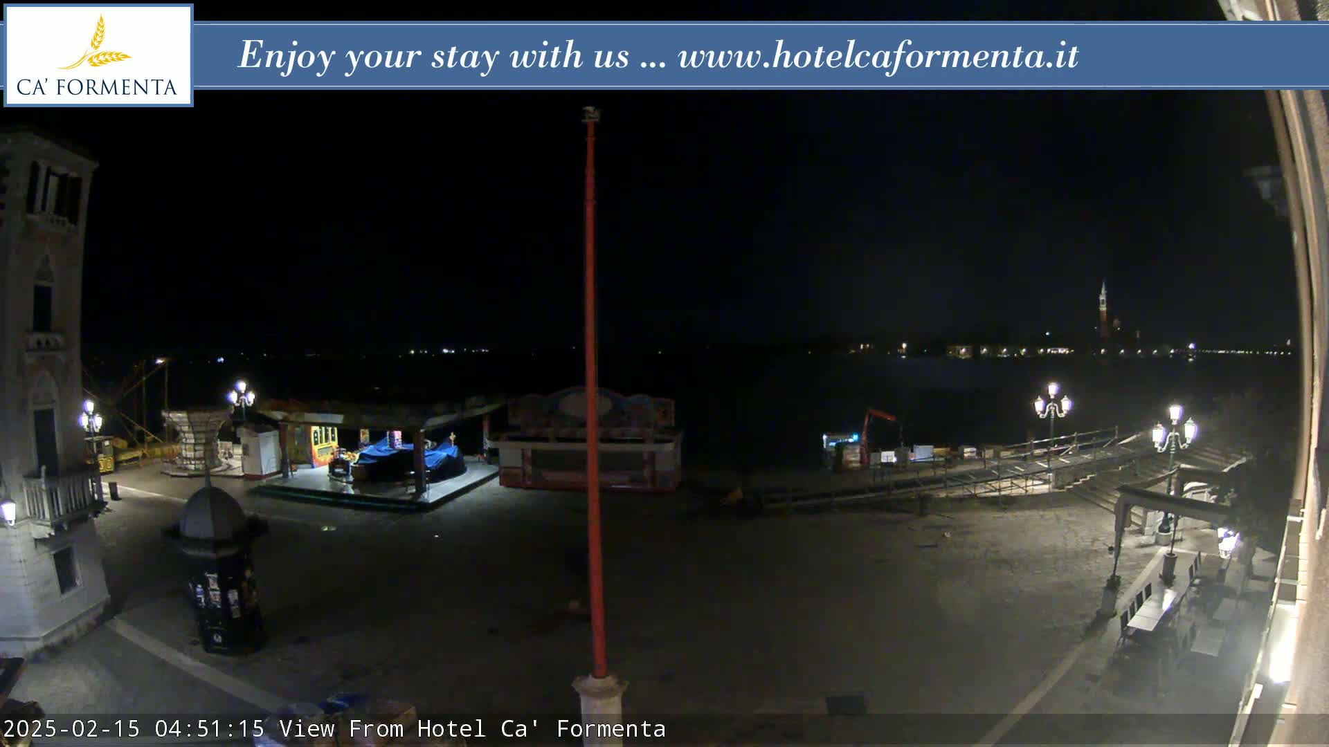 A nighttime view of a waterfront plaza, showing a mostly empty square with some small structures and distant lights across the water.