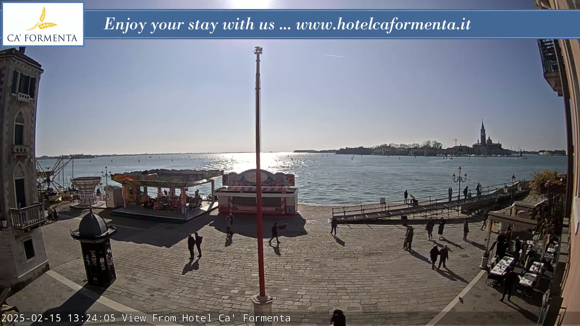 A sunny day reveals a waterfront plaza with a small amusement park, several people strolling, and a distant view of a city across the water.