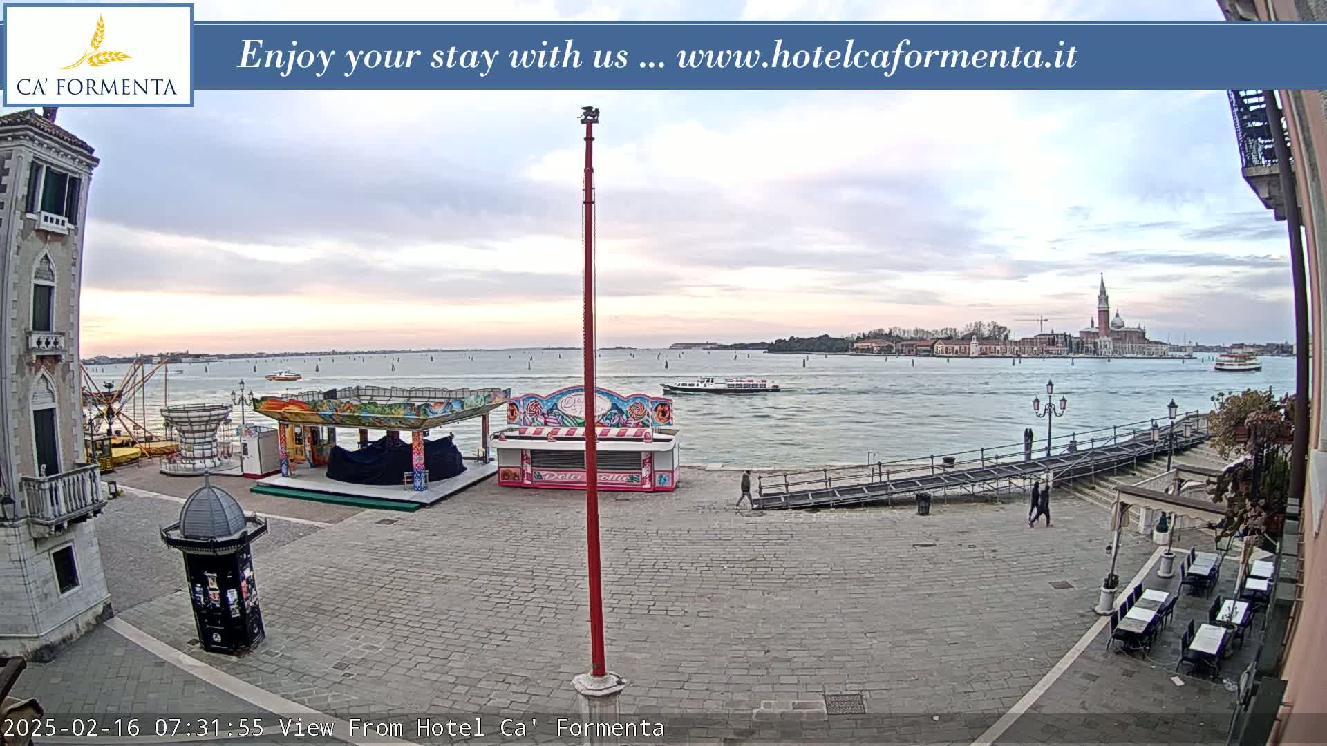 An overcast day reveals a waterfront plaza with a small carnival, a boat on the water, and a distant cityscape under a pale sky.