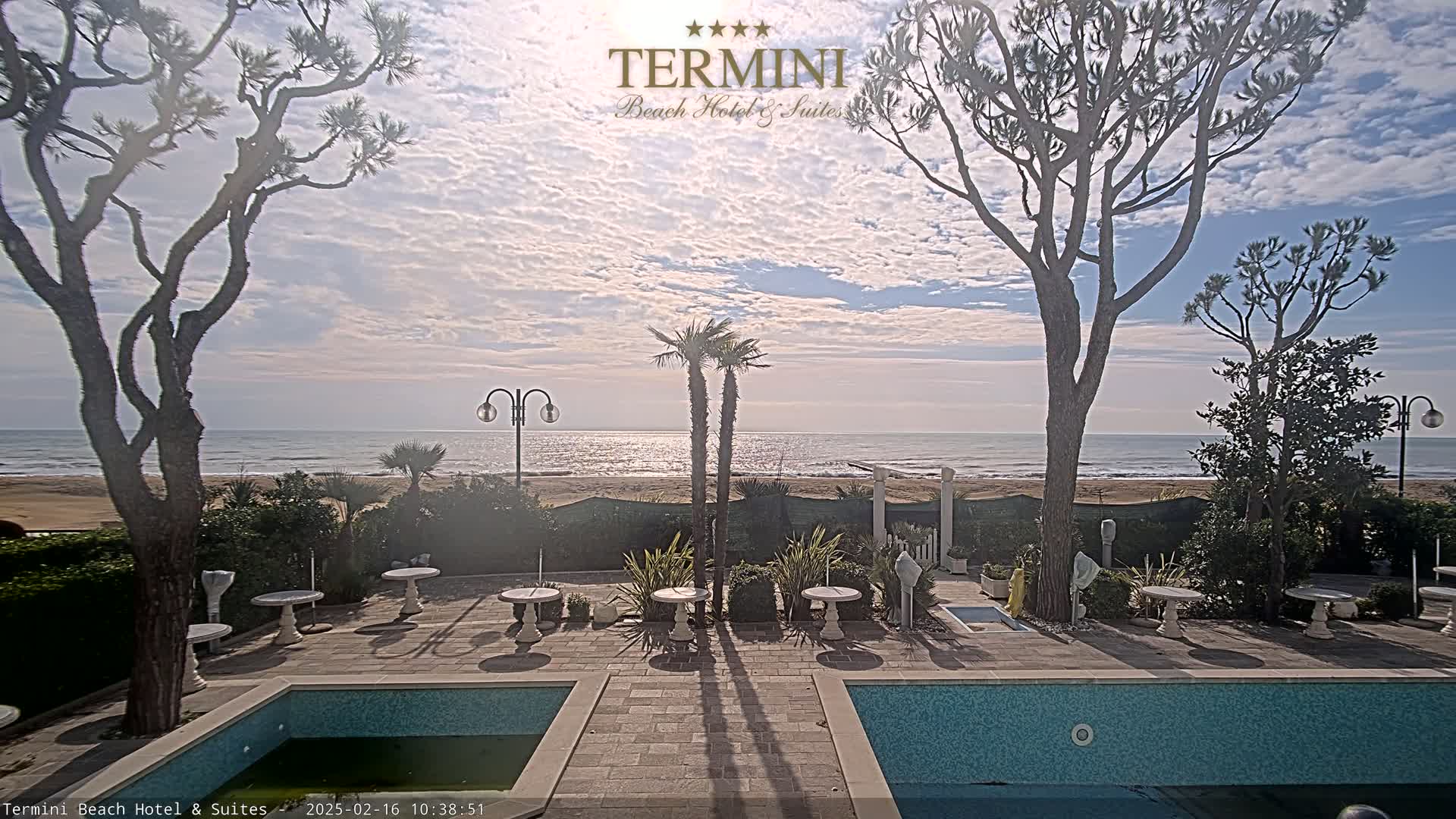 A partly cloudy day shows a tranquil ocean scene viewed from a patio with two swimming pools, surrounded by trees, plants, and small tables.