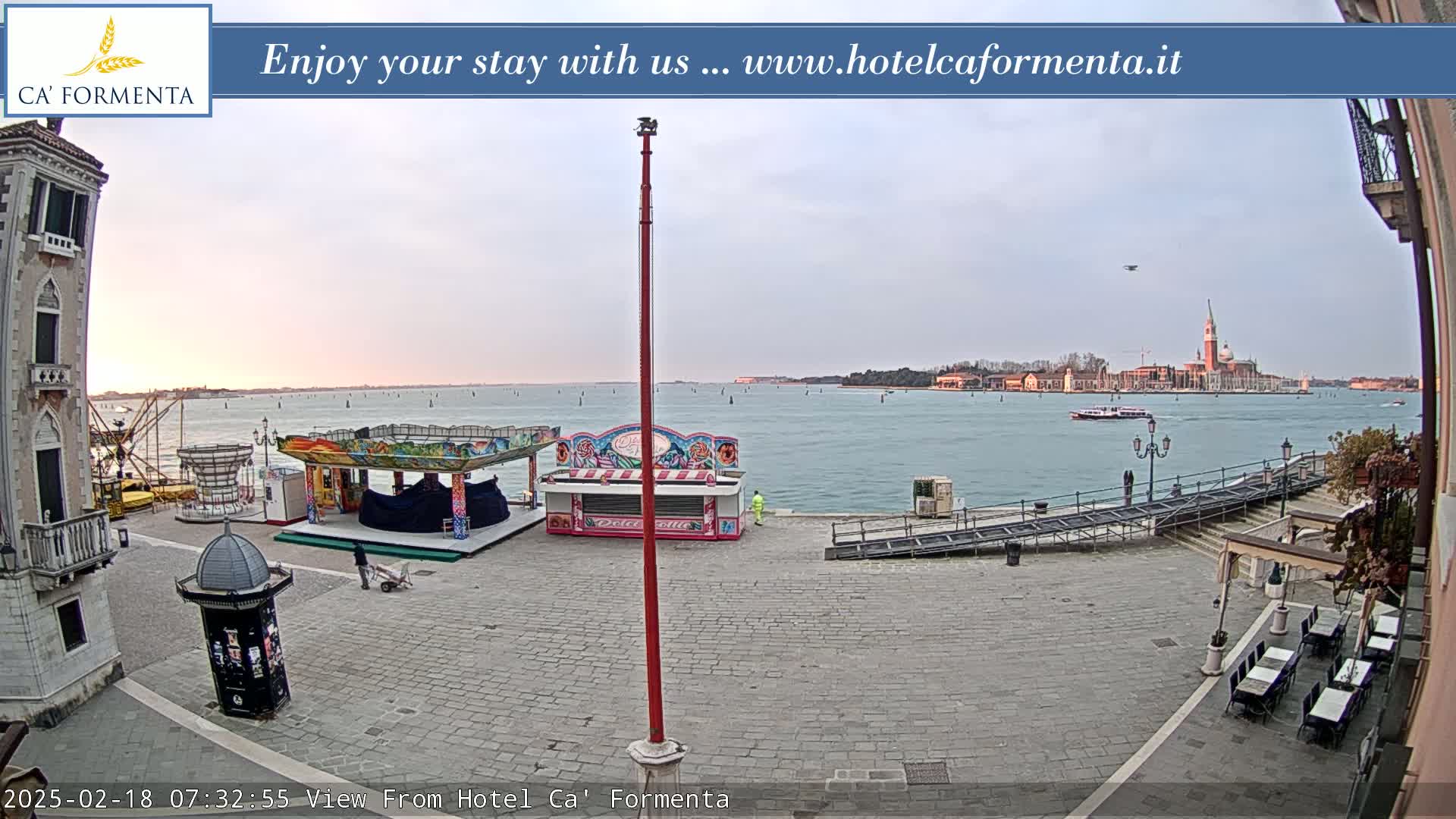 A waterfront plaza with a small fair and calm water showing a distant cityscape under an overcast sky.