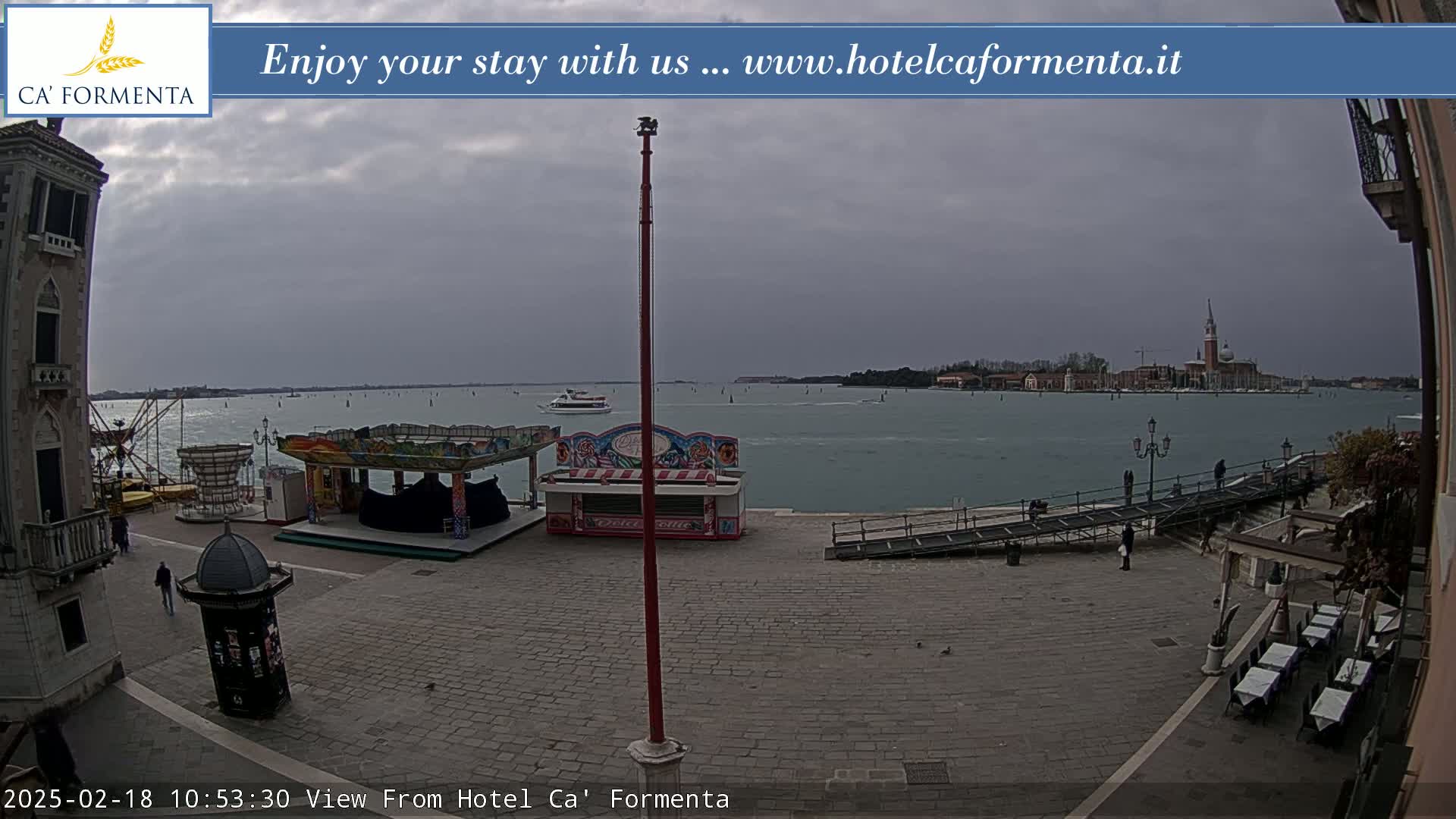 A waterfront plaza with a carousel and small kiosks, under an overcast sky, overlooks a calm body of water and a distant island.