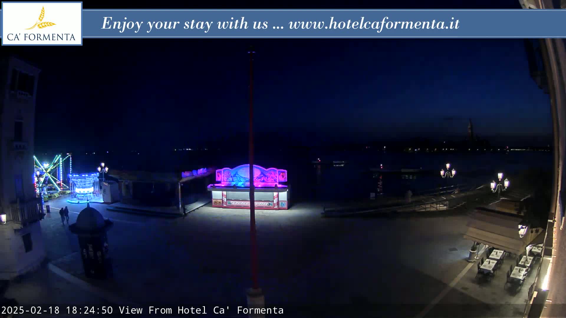 A nighttime outdoor scene shows a waterfront area with a few illuminated carnival rides, a brightly lit kiosk, and an outdoor restaurant with tables and chairs.