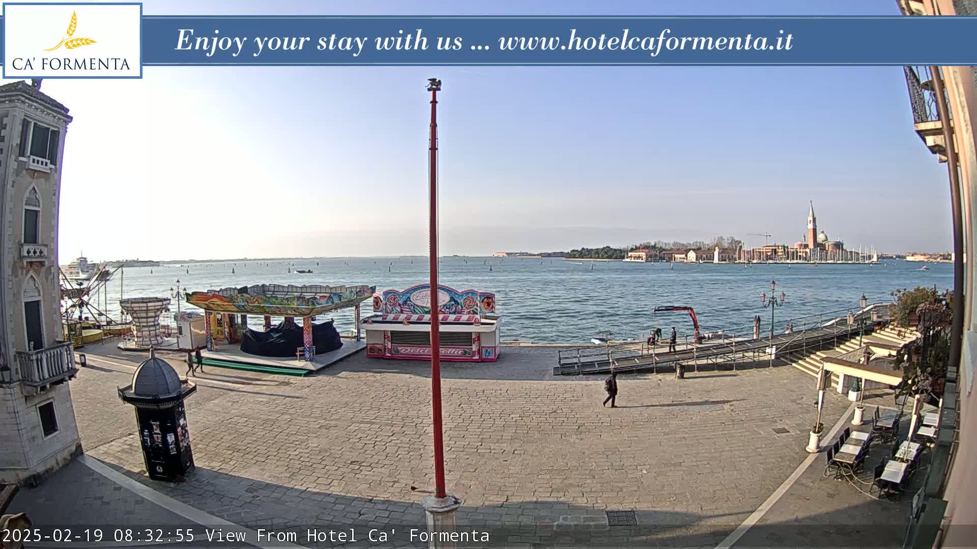 A waterfront plaza with a small amusement park and outdoor seating area overlooks a calm body of water under a clear, sunny sky.