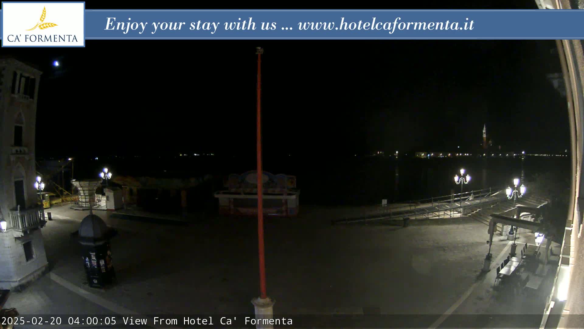 A nighttime view of a waterfront plaza with dimly lit buildings and a body of water showing distant lights.