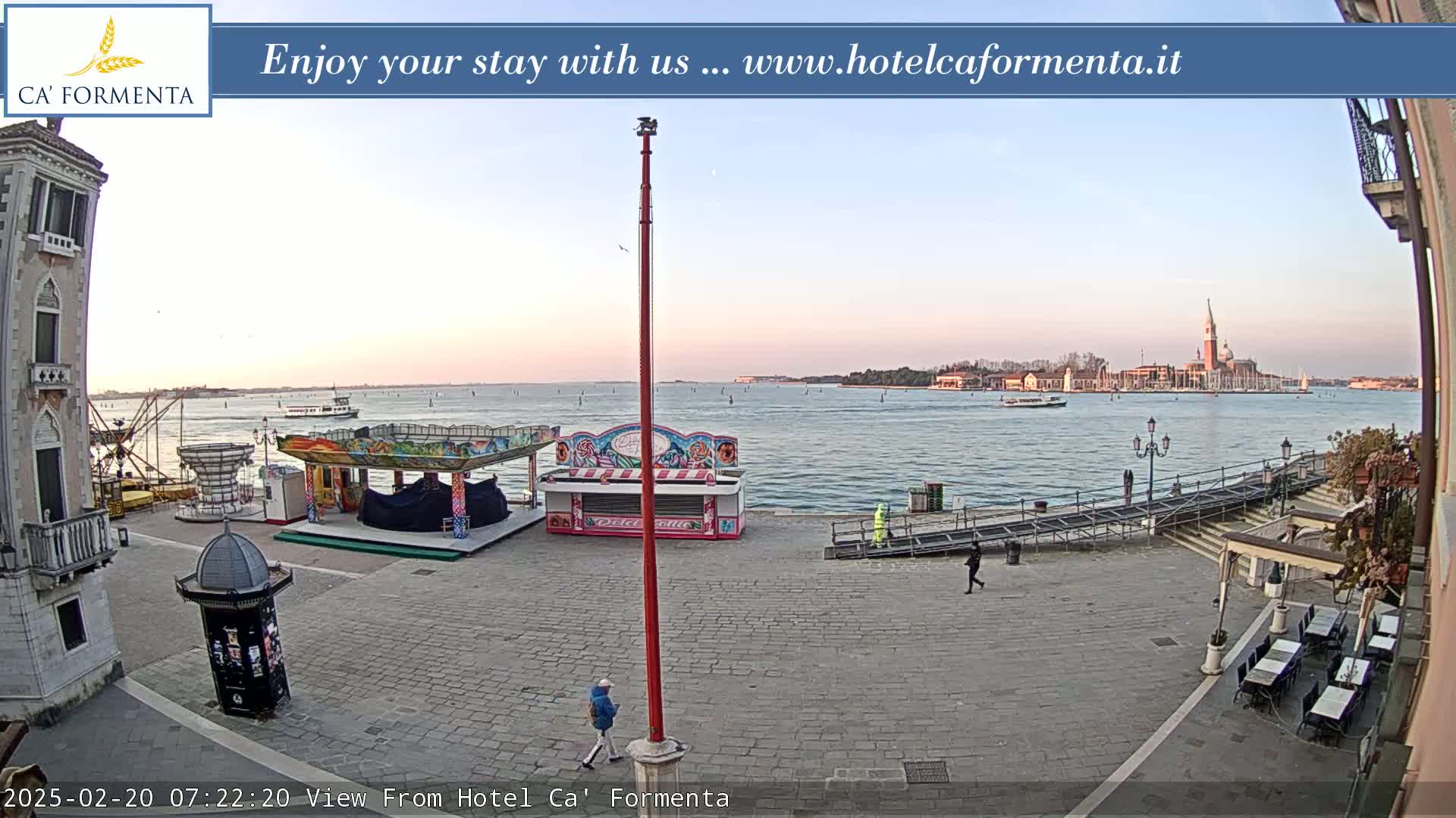 A calm waterfront square with carnival-style booths, a few people walking, and a distant view of a city across the water under a pale, clear sky.