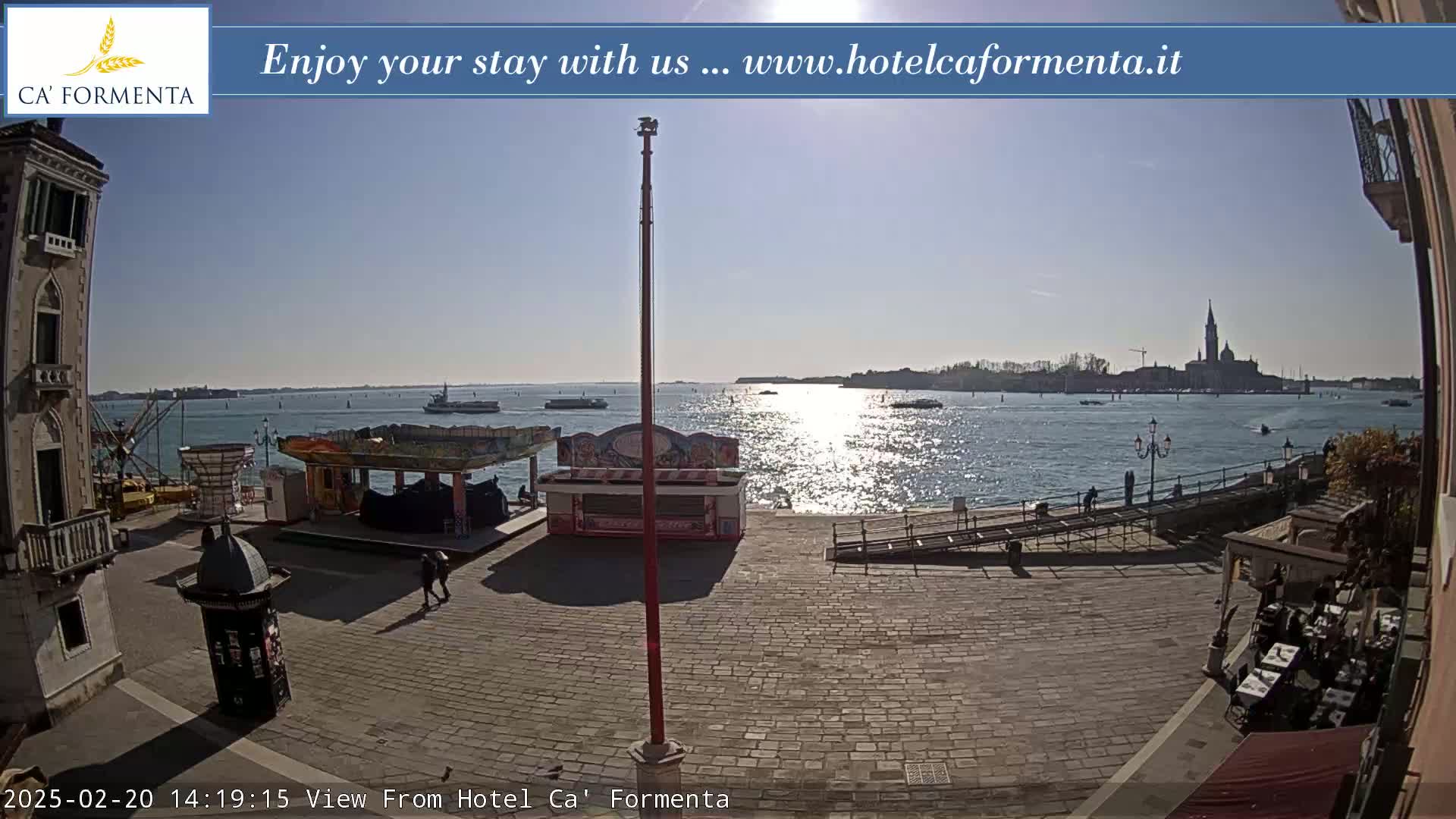 A sunny waterfront plaza with a few people, small kiosks, and a body of water showing boats and a distant cityscape.