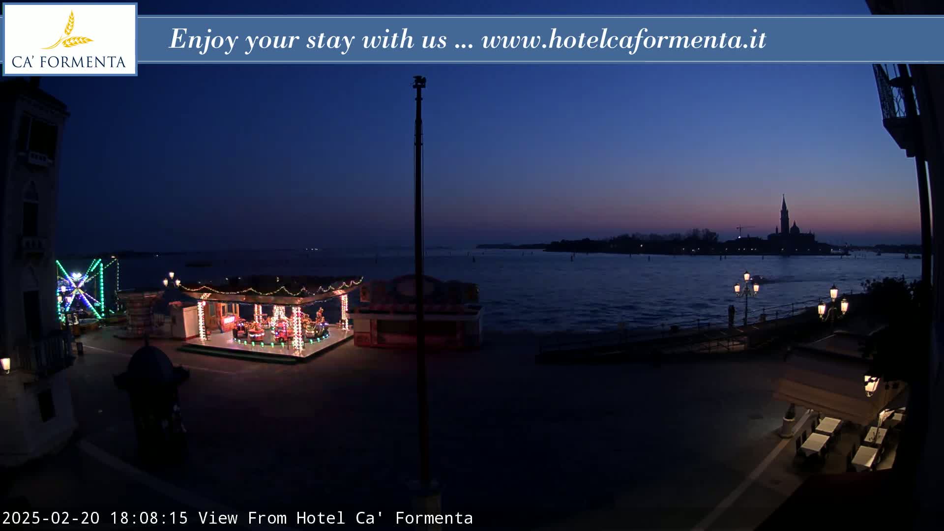 A nighttime view of a waterfront area shows illuminated carnival rides and a calm body of water with a distant island silhouette under a twilight sky.