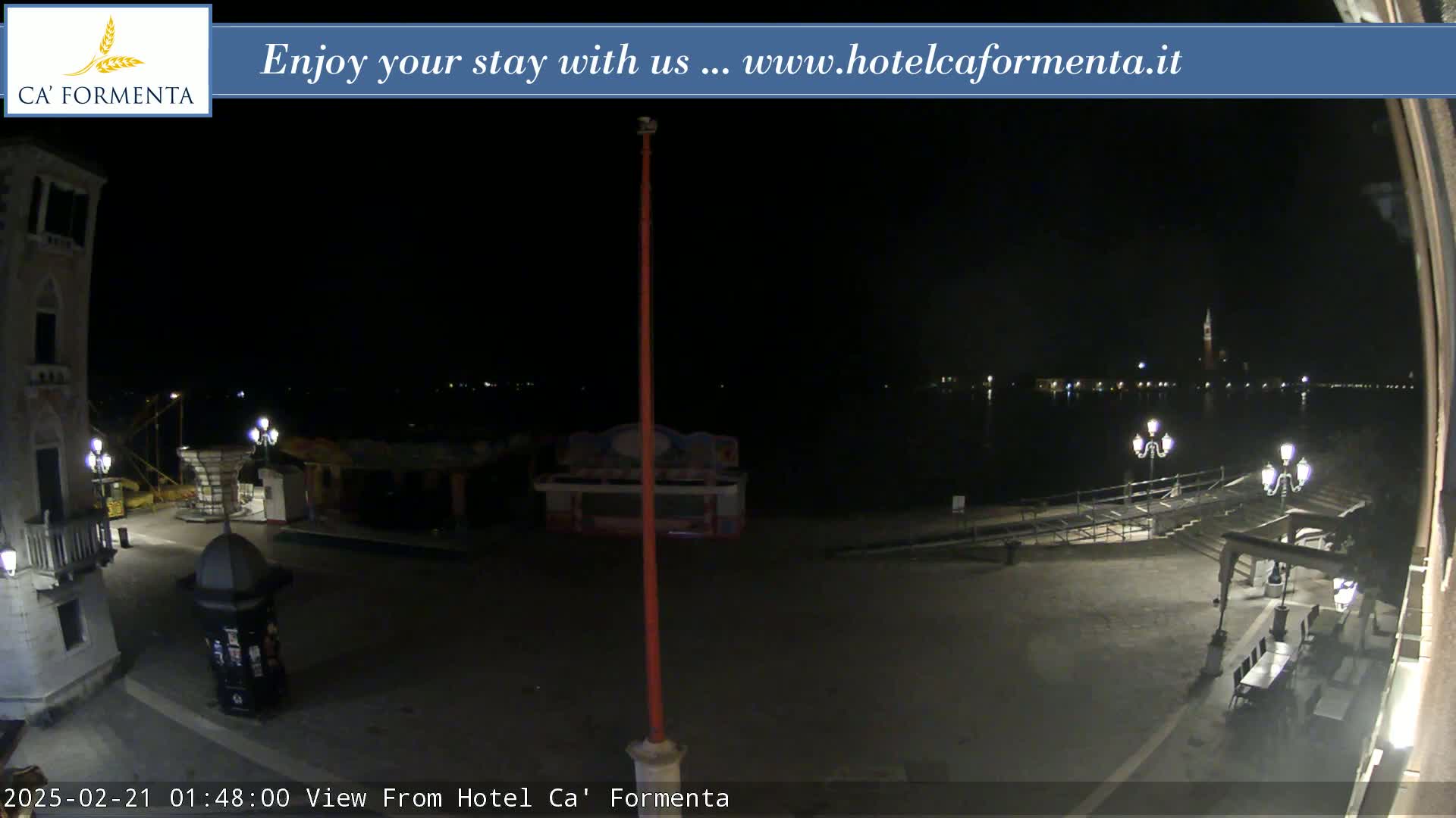 A nighttime view of a plaza and waterfront, with a calm body of water showing distant lights across it.