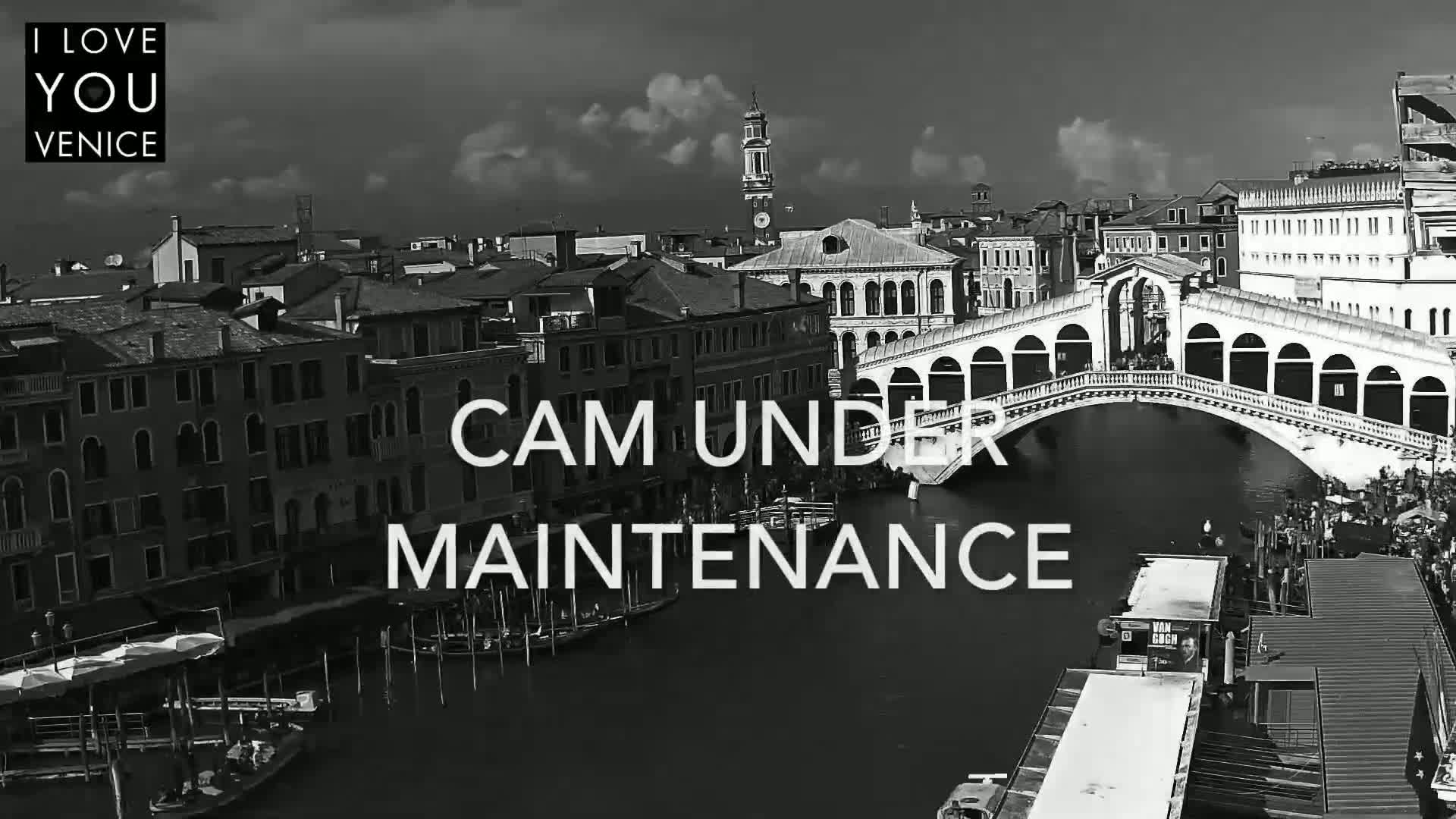 A black and white, high-angle view shows a canal in Venice, Italy, with boats and buildings along its banks, and a bridge spanning the waterway under a partly cloudy sky.