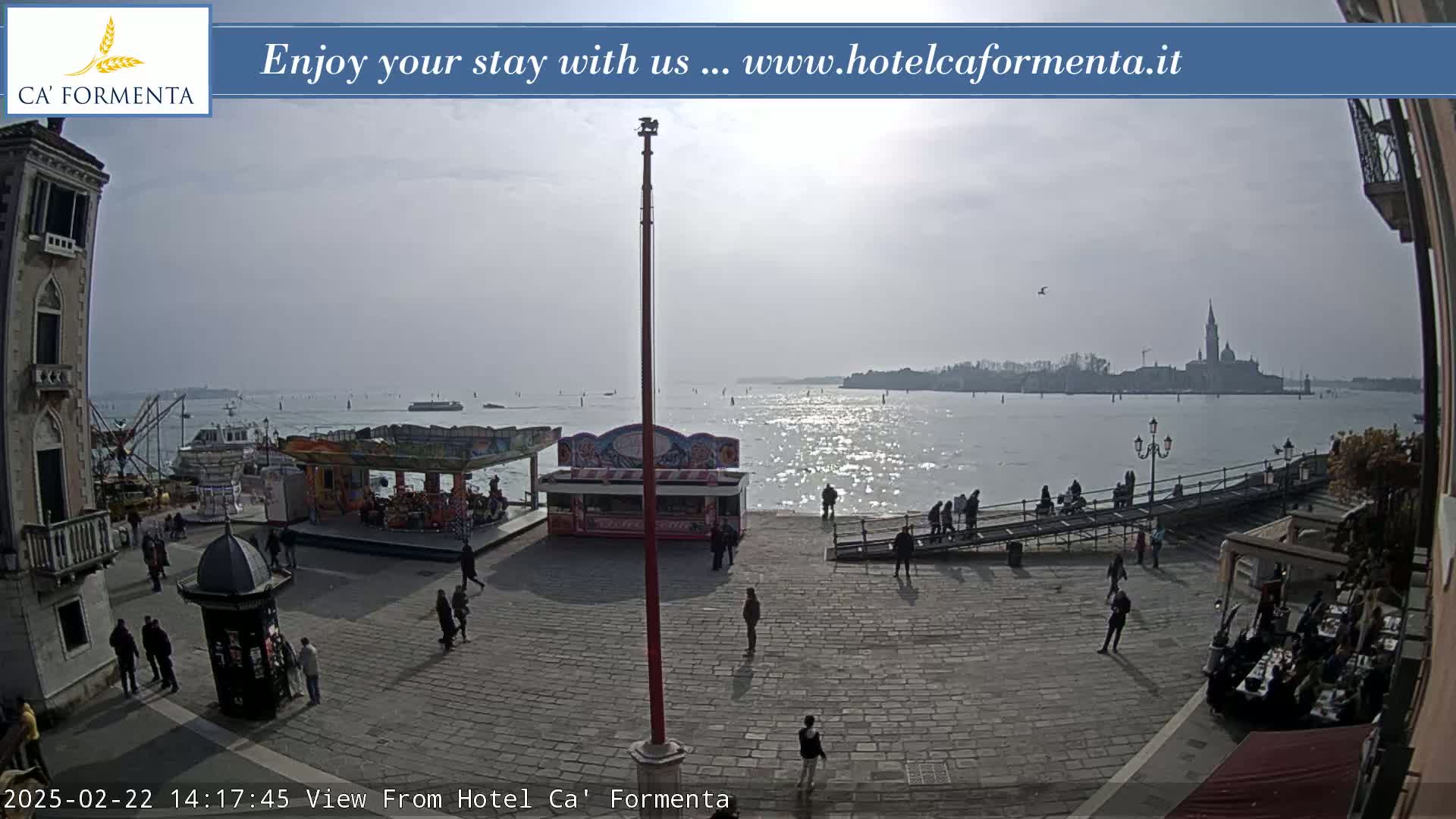 A sunny day shows a waterfront plaza with a few people, some small amusement park rides, and a distant view of a church across calm water.