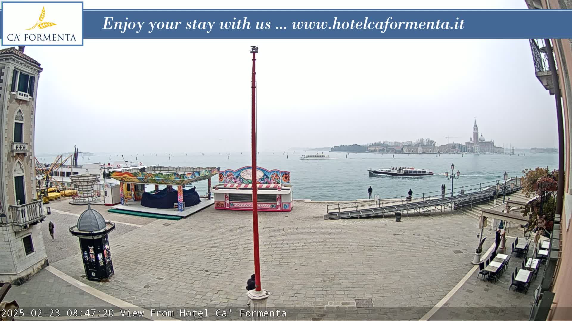 A paved waterfront square features a small fairground, with boats on the calm, overcast water beyond.