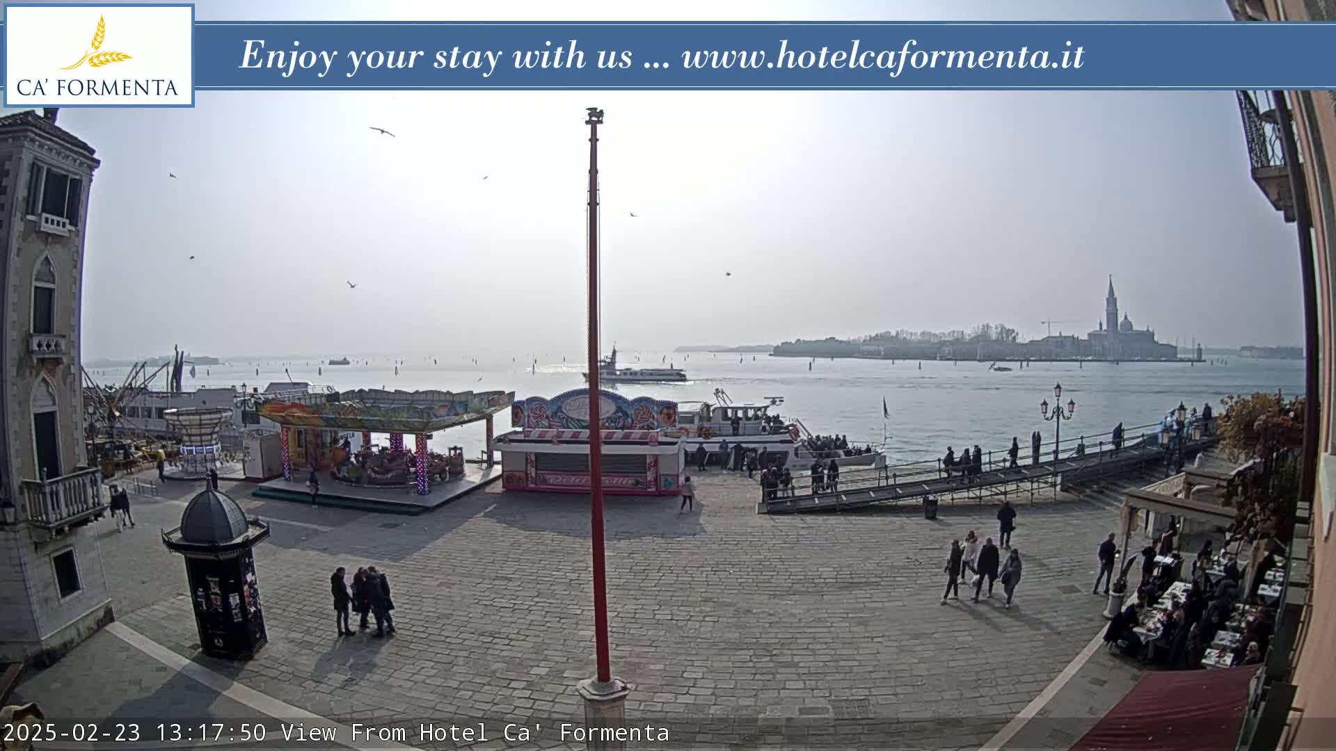 A paved plaza with a carousel and several small food stands overlooks a calm body of water with boats and a distant island under a hazy, sunny sky.
