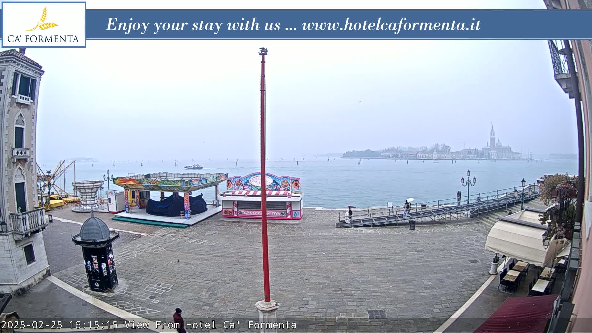 A hazy, overcast view of a waterfront plaza with carnival booths, a stone walkway, and a distant island.