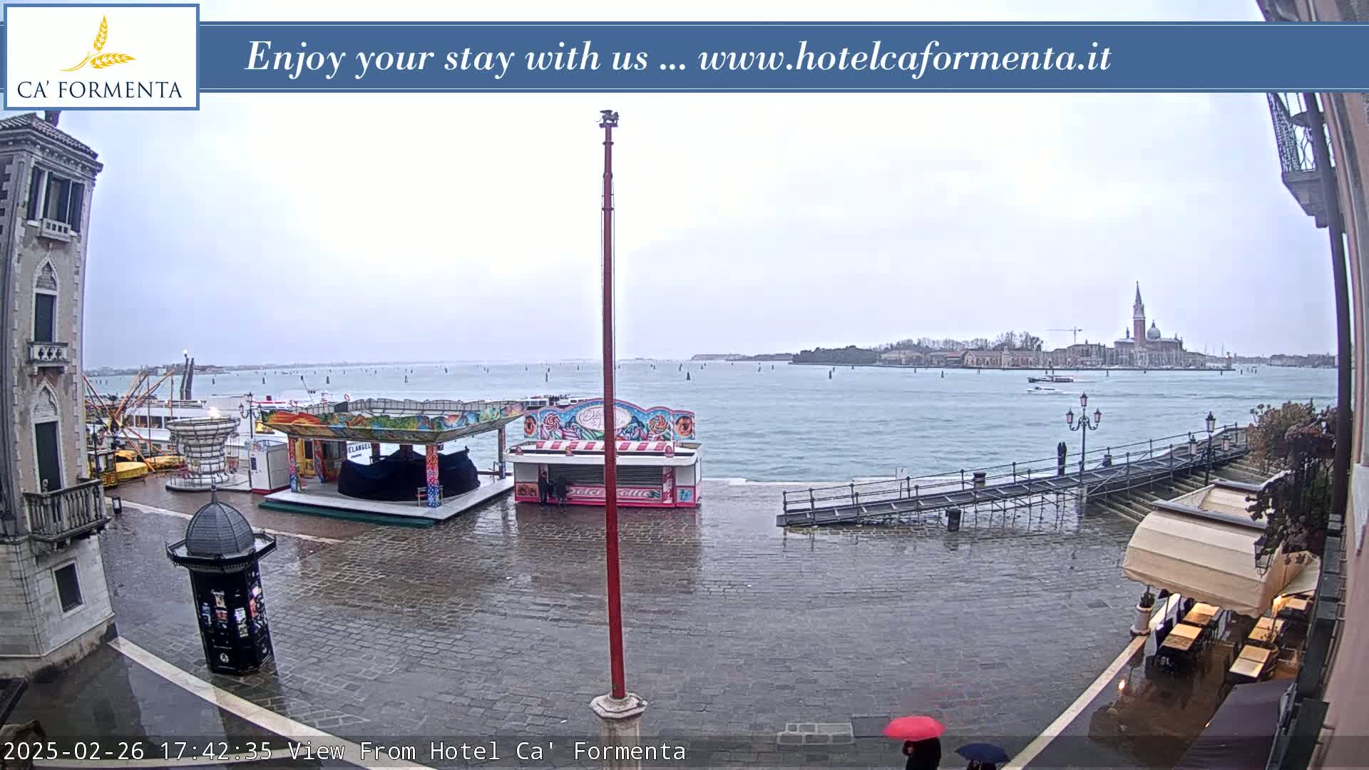 A rain-soaked waterfront plaza shows a few carnival-style food stands and a distant view of a city across a body of water.