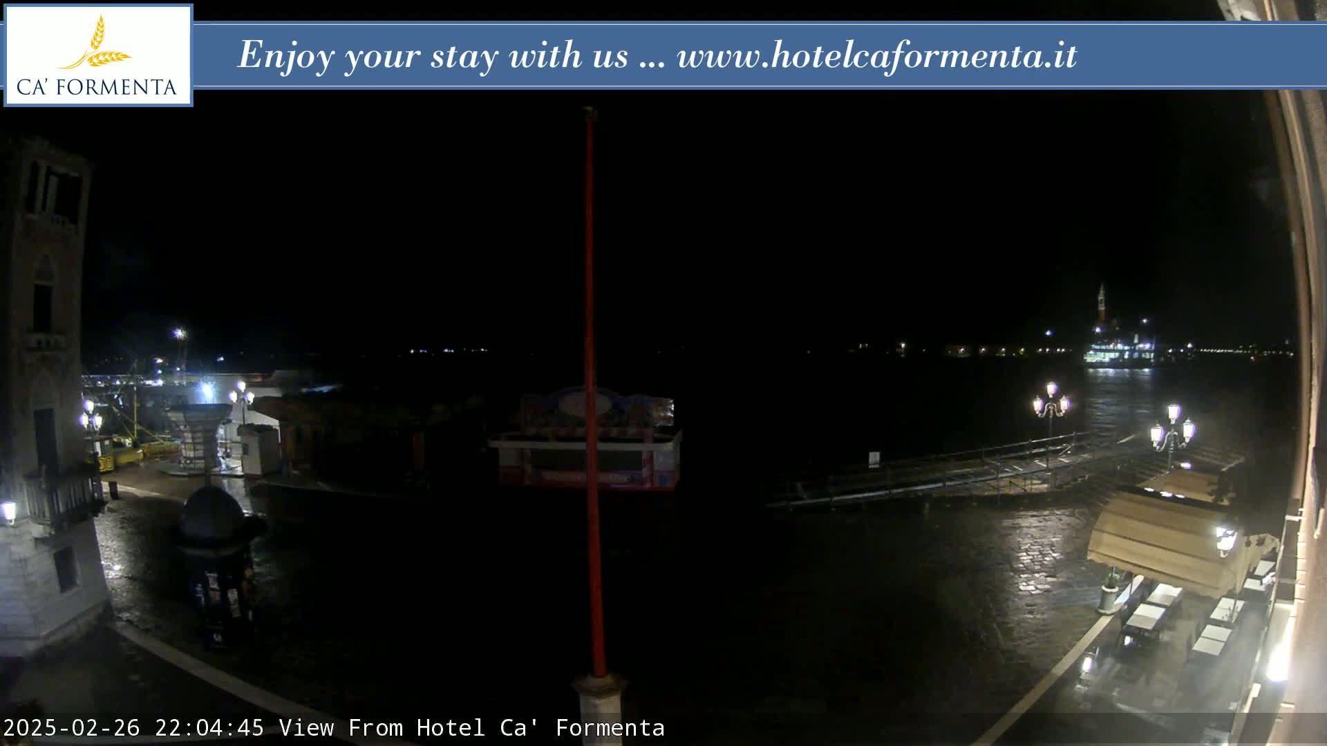 A nighttime view of a waterfront plaza, partially submerged in water, with a building and distant lights across a body of water.
