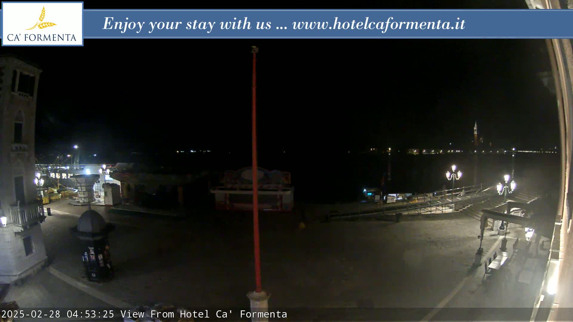 A nighttime view of a waterfront plaza with a calm body of water and distant lights across it.
