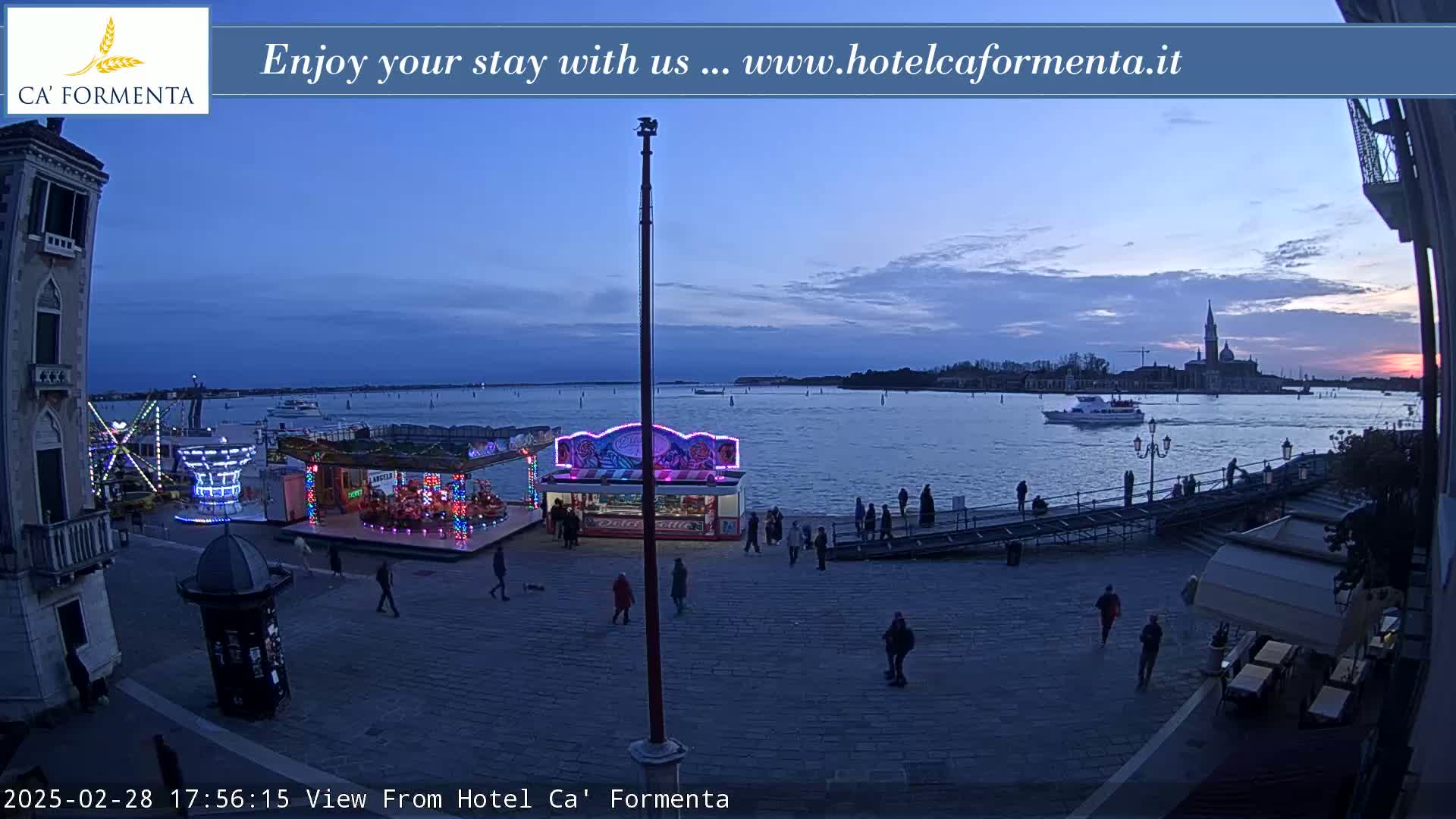 A twilight view shows a waterfront plaza with a small amusement park, a few people, and a boat on the water near a distant church.