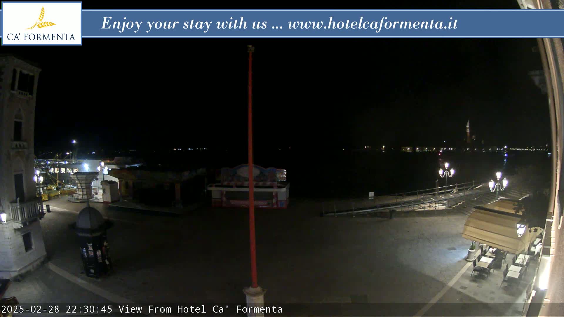 A nighttime view of a waterfront plaza shows a calm body of water, some outdoor seating at a restaurant, and a few carnival-like structures.