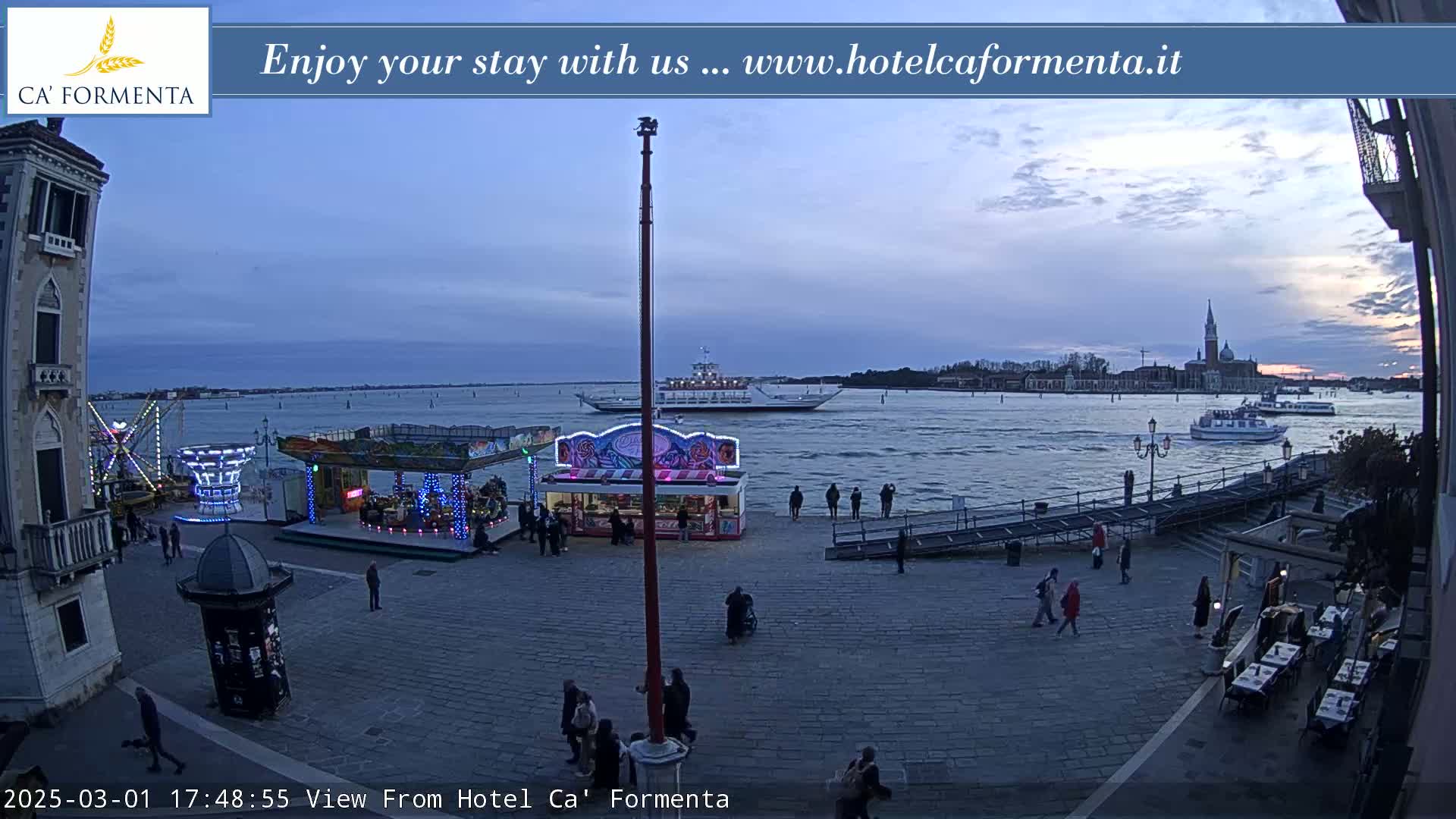 A waterfront plaza with carnival rides and a few people milling about, overlooks a body of water where boats are docked under a twilight sky.