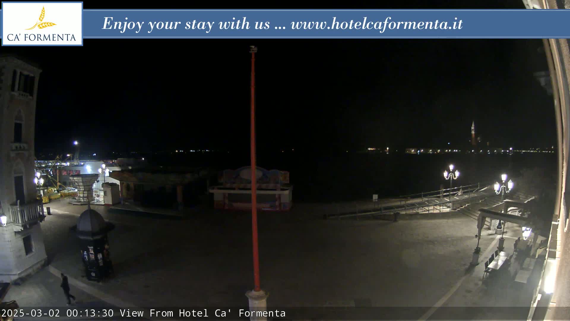 A nighttime view of a waterfront plaza with a few lights, some small structures, and a body of water with distant lights across it.