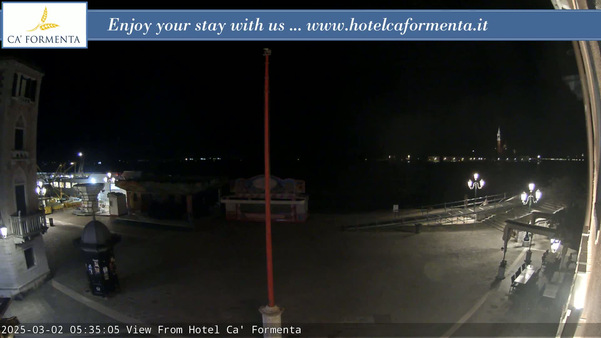 A nighttime view of a waterfront plaza with a few lights, some small structures, and a body of water with distant lights in the background.