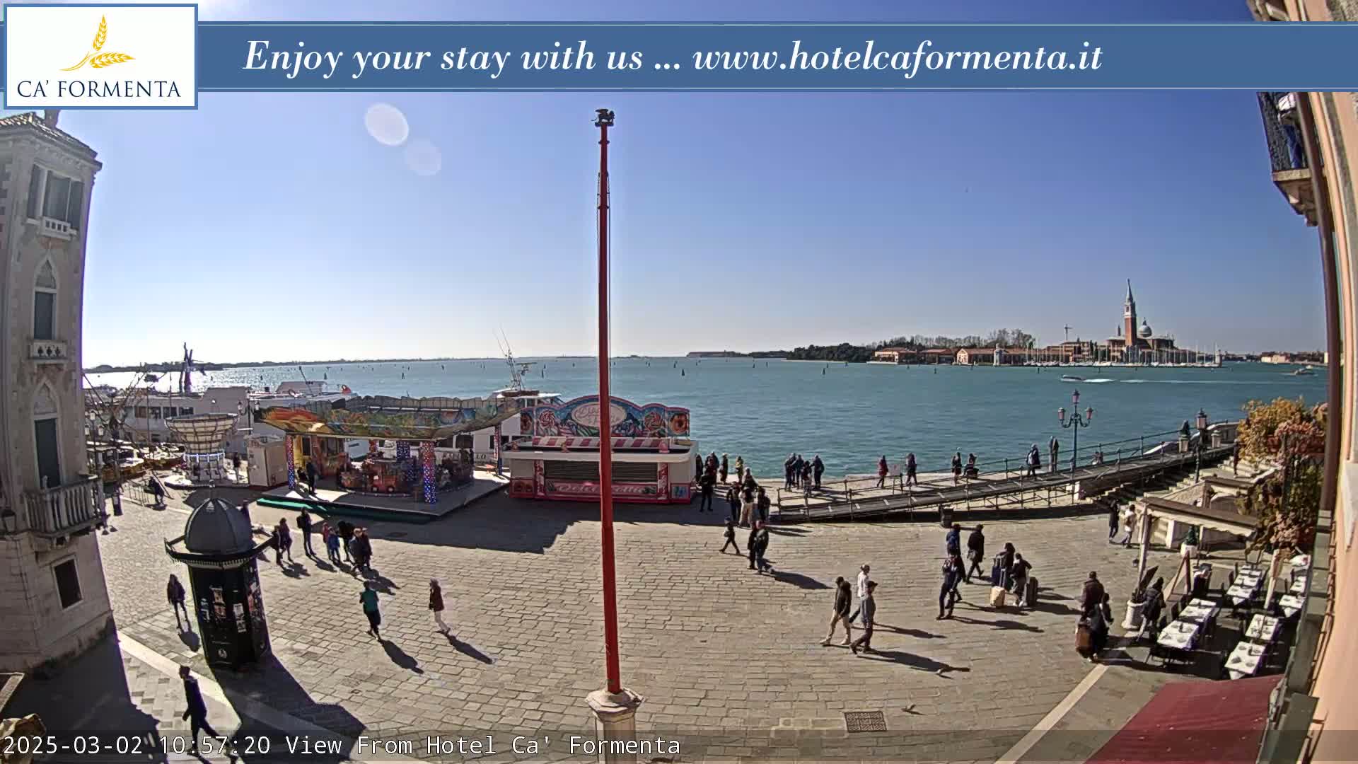 A sunny outdoor scene shows a paved plaza with a few people walking around, near a body of water with boats and a distant island, and carnival-style booths and rides.
