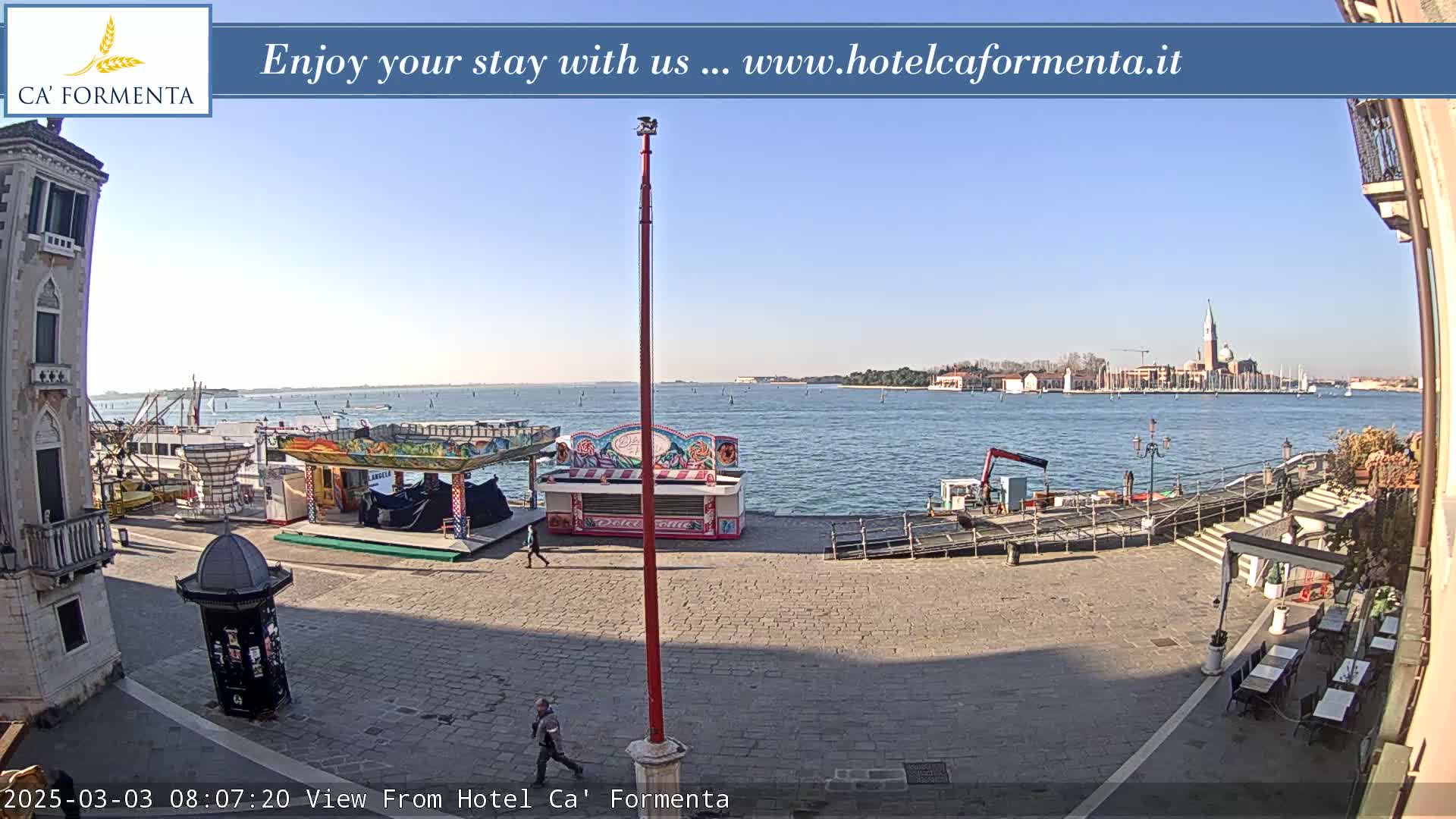 A sunny waterfront plaza with a few people, small carnival-style stands, and a view of a distant island across calm water.