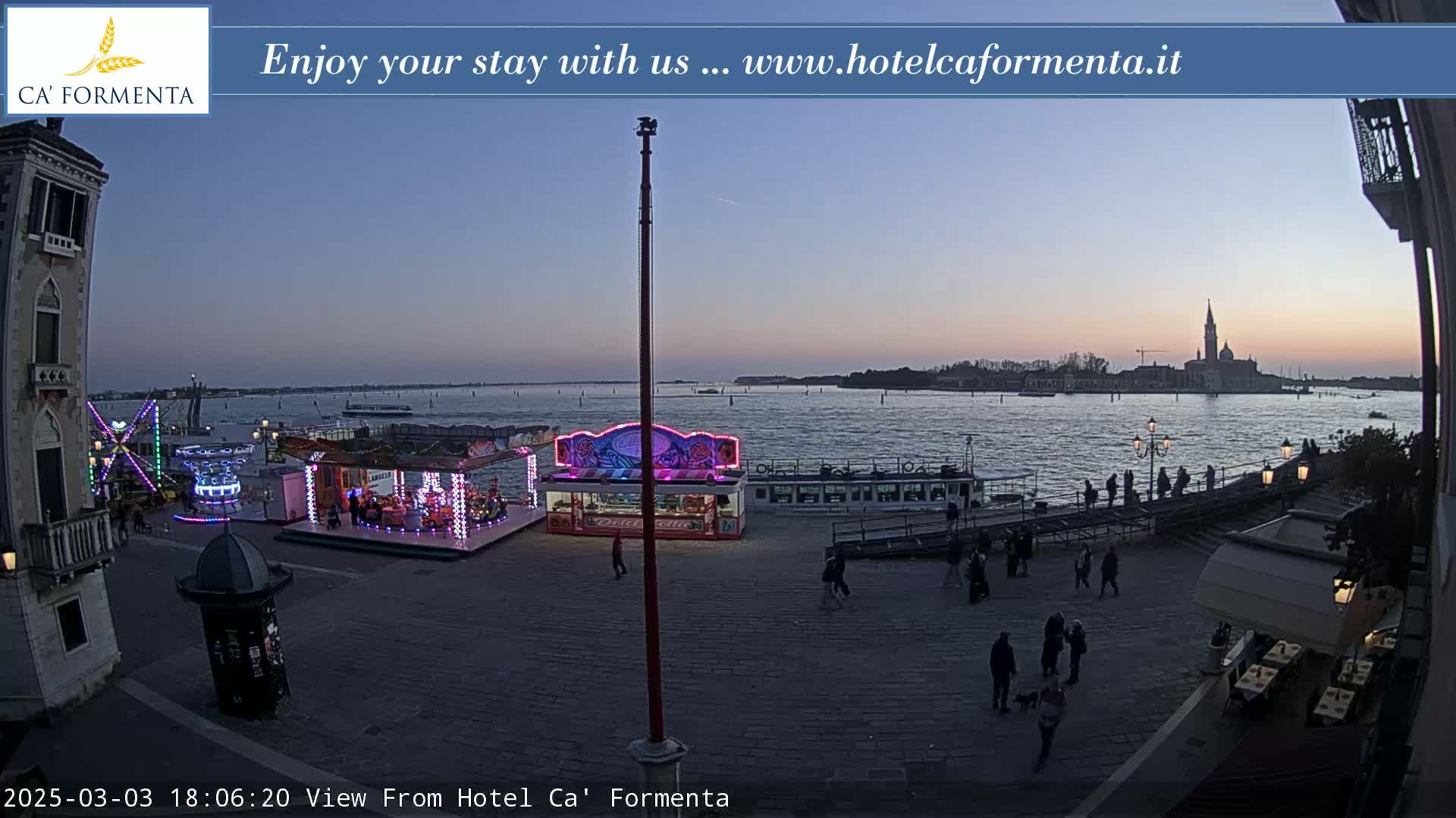 A twilight view shows a waterfront plaza with carnival rides, a restaurant, and people walking, overlooking a calm body of water and distant buildings under a dusky sky.