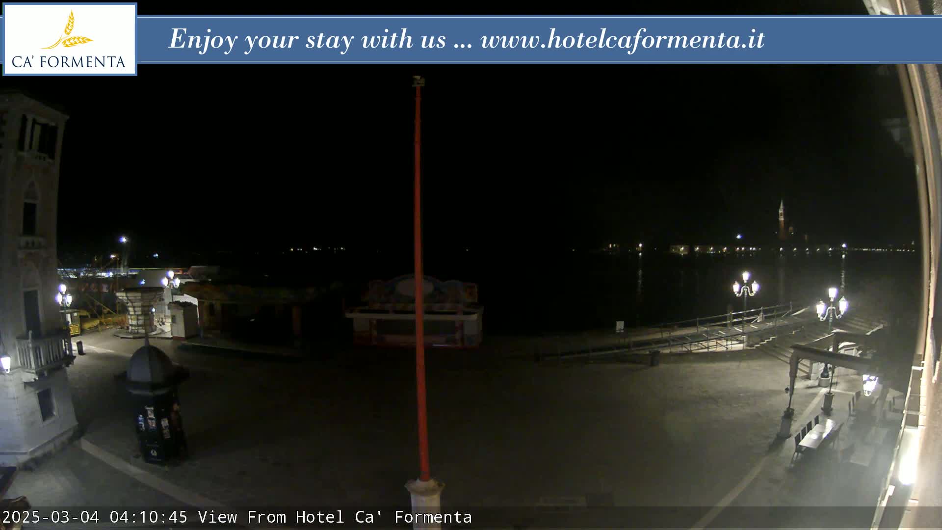 A nighttime view of a waterfront plaza with a calm body of water and distant lights across it.