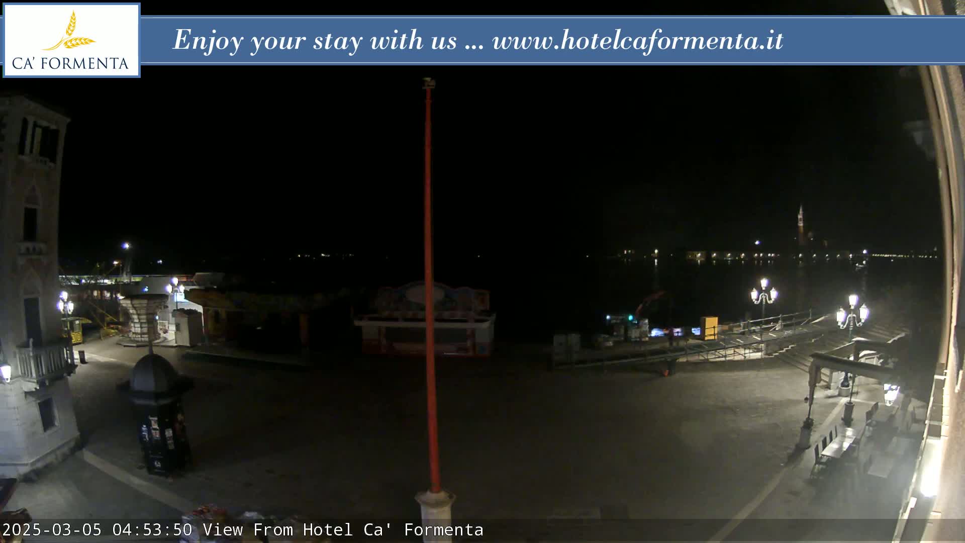 A nighttime view of a waterfront plaza with a few lights and some closed kiosks, overlooking calm dark water and distant city lights.